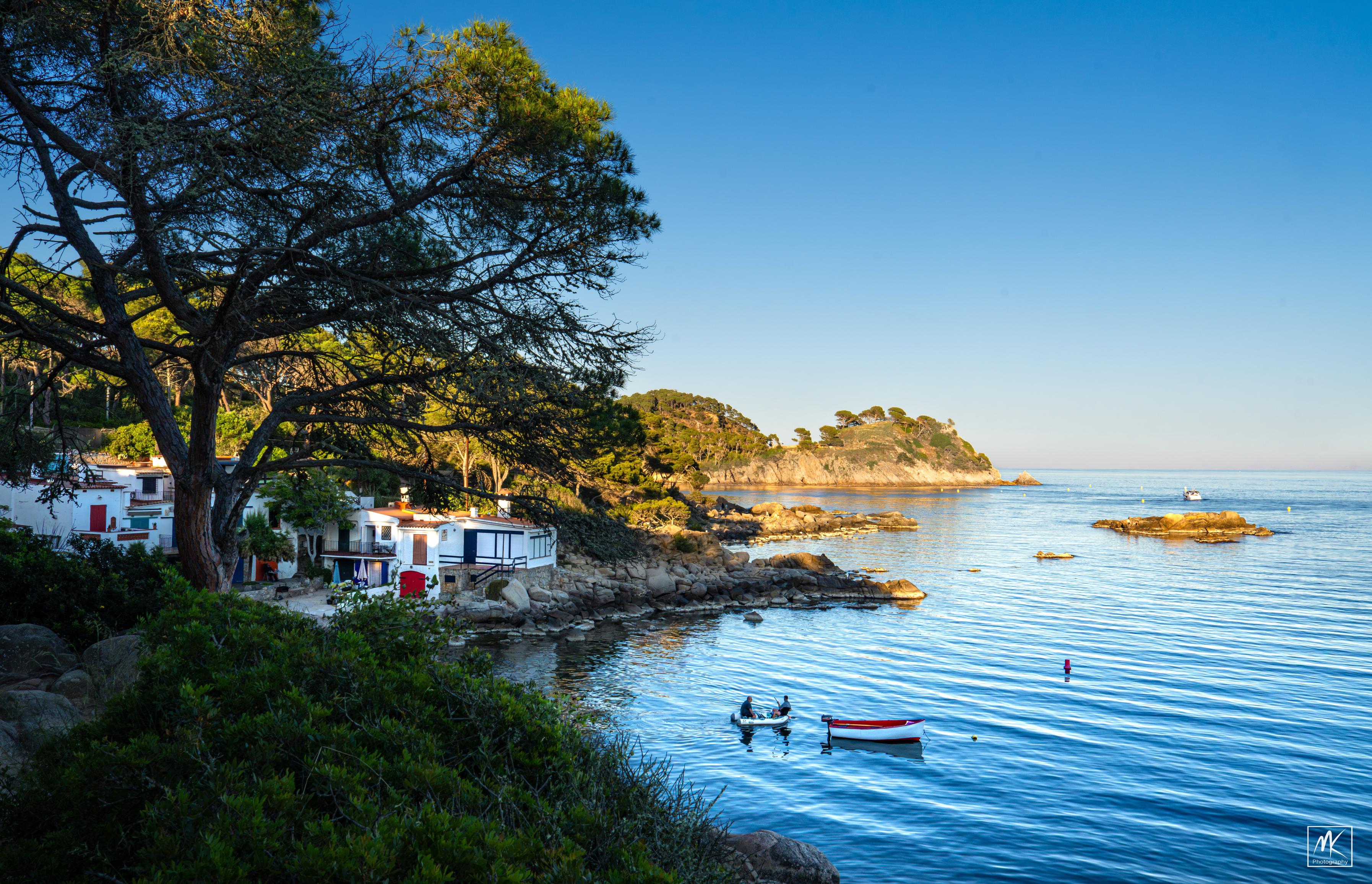 Color photo of a rocky seashore with small coves and islets in the water. In the foreground is a large tree and beyond are the white buildings of a small fishing village. In the water in the foreground are two people in a tiny inflatable skiff approaching a small wooden fishing boat that is moored offshore.
