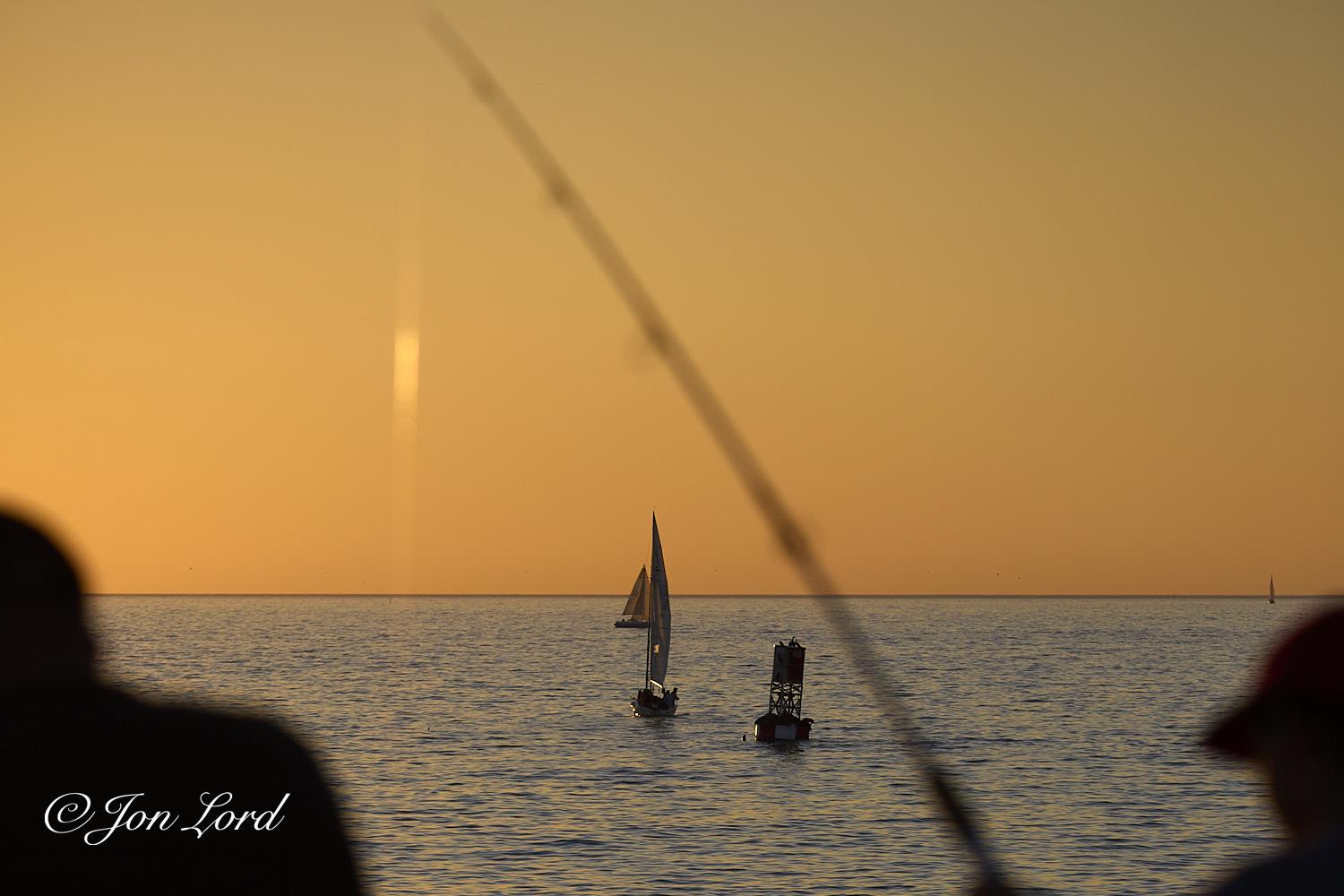 This is a colour sunset photo in landscape format of two people fishing in the sea from a pier in the foreground, three sailing boats and a buoy in the middle ground and a golden sunset in the background. Redondo Beach, California (2013).

In the lower left and right corners are the silhouettes of the two people fishing. The person on the right is holding their fishing rod that extends diagonally up and across, passing near the centre of the photo and with the fishing line hanging vertically to the left of centre and glowing from the light of the setting sun. In the middle ground are the blue waters of the Pacific Ocean with small waves disturbing the surface. Below the centre and slightly to the right, fifteen metres away, is a large metal navigation buoy with at least one large seal or sea lion on the right side causing the buoy to tilt to the right. Below centre is the silhouette of a sailing boat, underway and sailing away from the camera, a short distance beyond the buoy. Beyond t
