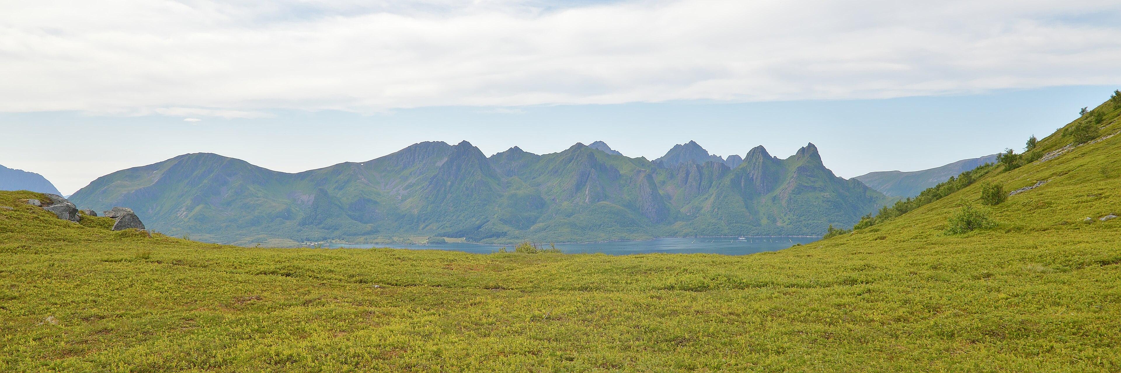 A panoramic photo looking off the green side of a mountain across to a mountainous island. The sky is hazy with clouds in it.