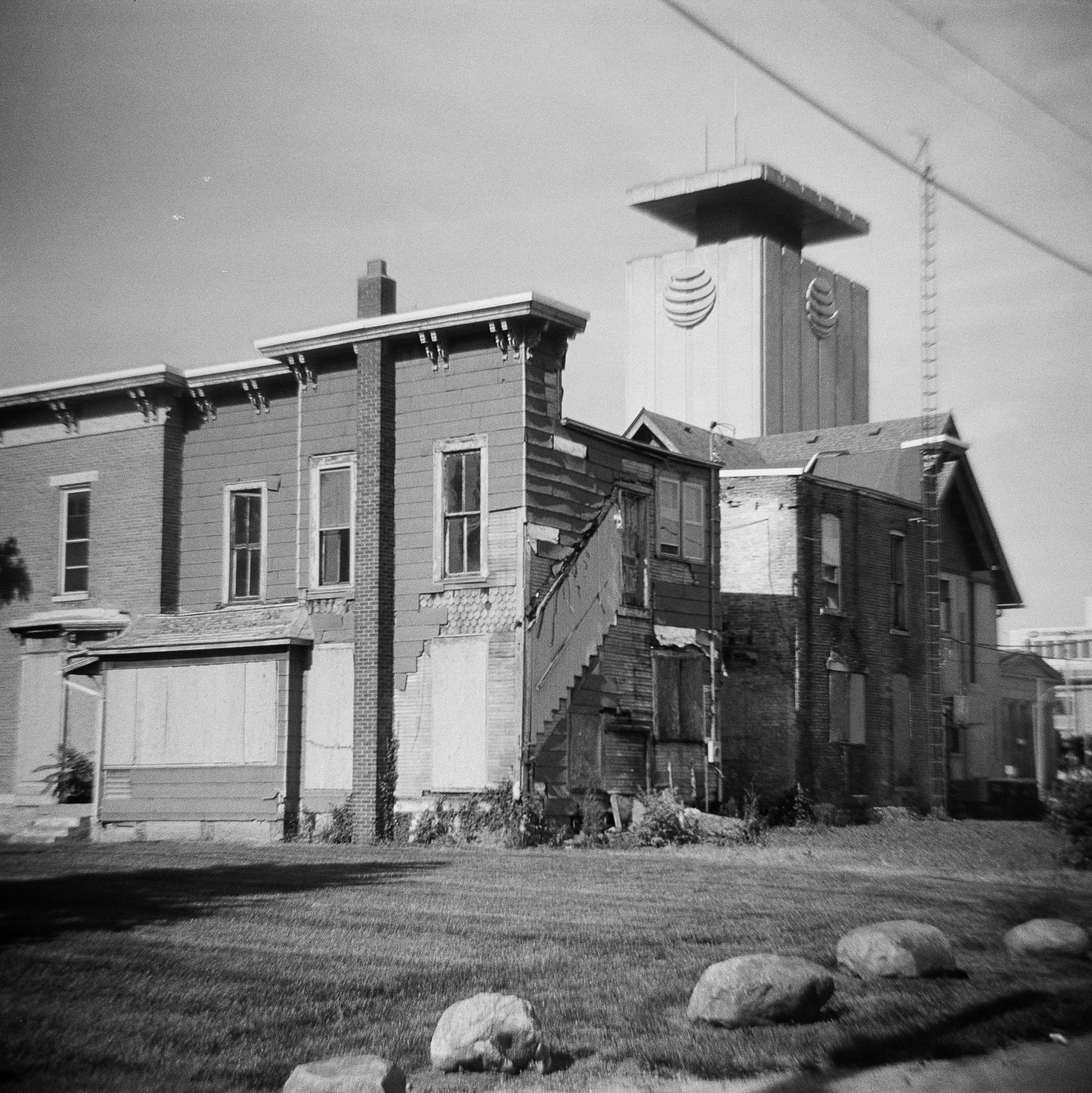 A row of buildings, apparently very old homes, viewed from the back. The roof of the closest building has trim at the top that is ornate, in a style that appears to be from the 1800s. The windows are narrow and tall. There is one tall, narrow chimney extending from the ground to beyond the top of the building. The lower windows are boarded up, and siding appears to have fallen off of the building. In the back, it looks like there was once a stairway on the exterior of the building, but now it is gone. The buildings beyond this one also appear to be in disrepair. A tall tower for a TV antenna is seen attached to one of the buildings. In the background, a much taller building is seen rising above the closer buildings. It appears to be made of concrete, with vertical lines on the surface. Near the top we can see two large AT&T logos. At the top of the building, the structure narrows, but there is a wider, flat roof at the top. In the upper right corner of the frame we see some electrical 