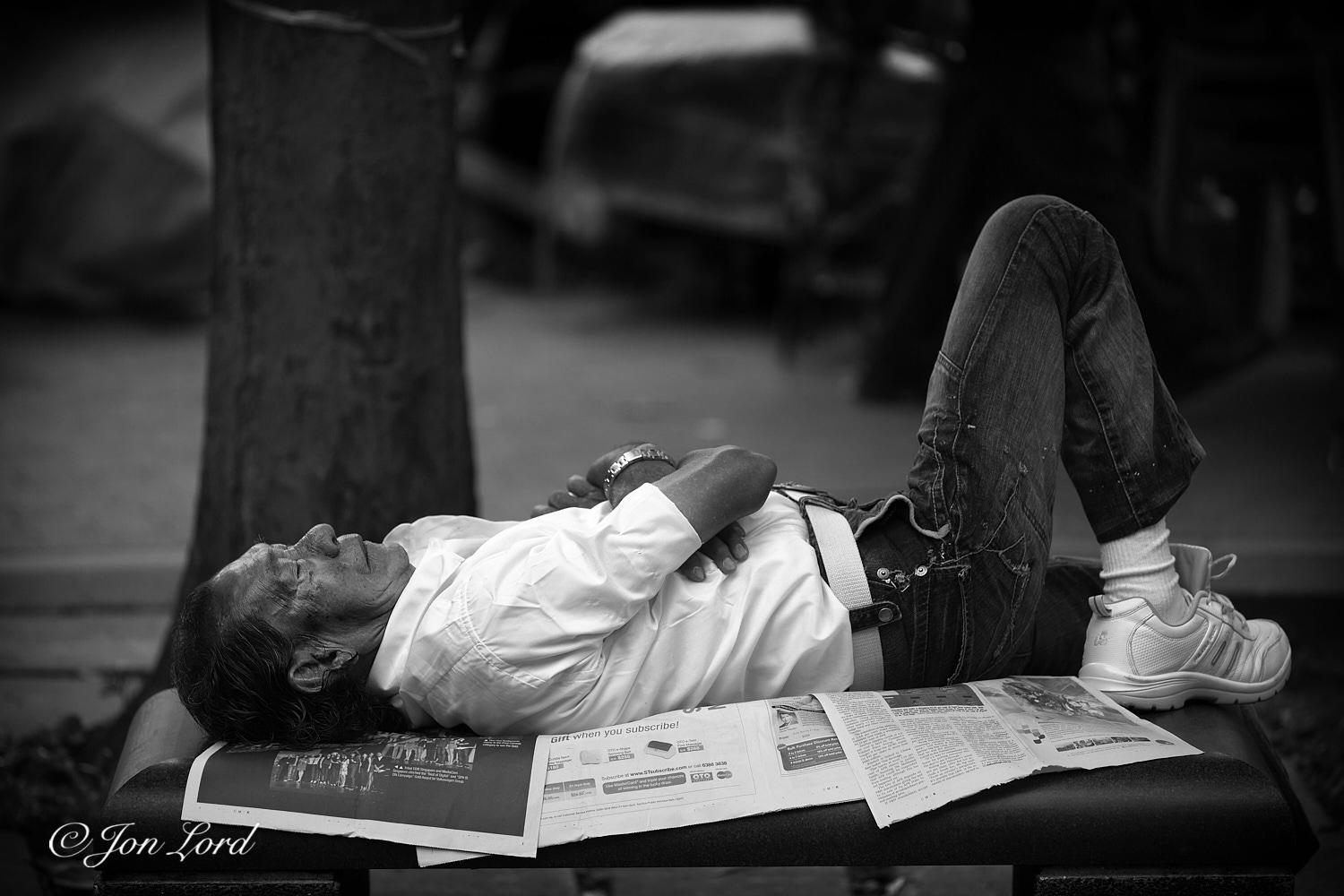 This is a black and white street photo in landscape format of a middle aged + local man, lying asleep on a park bench. Rochor, Singapore (2013).

Below the centre and centred in the photo is our sleeping man. The camera is slightly above the level of the park bench and about two metres away. Our slumbering man is viewed in profile is lying face upwards, head to the left, feet to the right and is filling most of the length of the image. He is lying on three pages  of broadsheet newspaper. He has neat dark hair combed back, is wearing a white, short-sleeved shirt, denim trousers, white trainers and matching white socks. His arms are crossed over his stomach, while his right leg is elevated in an inverted 'V', his left leg is out of view, perhaps resting on the ground? The park bench does not have a back rest. Behind our man's head is the trunk of a medium sized tree. In the background, about five metres away, is a row of market stalls, closed and covered up: all heavily blurred out of fo
