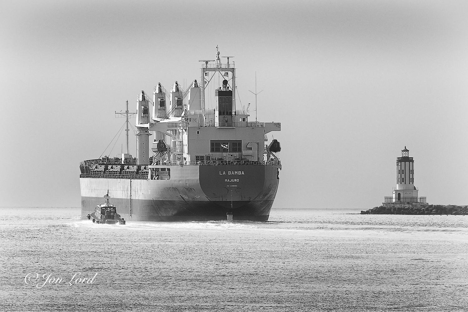 This is a black and white photo in landscape format of a Bulk Carrier ship leaving port and passing a lighthouse located at the end of a breakwater. San Pedro, California (2018).

The rippled and silver toned sea water rises from the base of the photo upwards to the one third up level. In the centre of the image is the stern of our medium sized (178m long) bulk cargo ship, underway and moving away from the camera at a slow speed, leaving little wake behind. The vessel is viewed obliquely with the stern and port side in view, with the ship filling a third of the width and a third of the height of the photo and viewed from about 20m away. The ship is riding high in the water with 3m of dark antifouling in view with a lighter toned hull above. The white accommodation and wheelhouse superstructure is close to the stern with a tall rectangular funnel above. Forward are four large cranes spaced evenly on the centreline of the deck. On the stern is the ships name 'LA BAMBA' in large white let