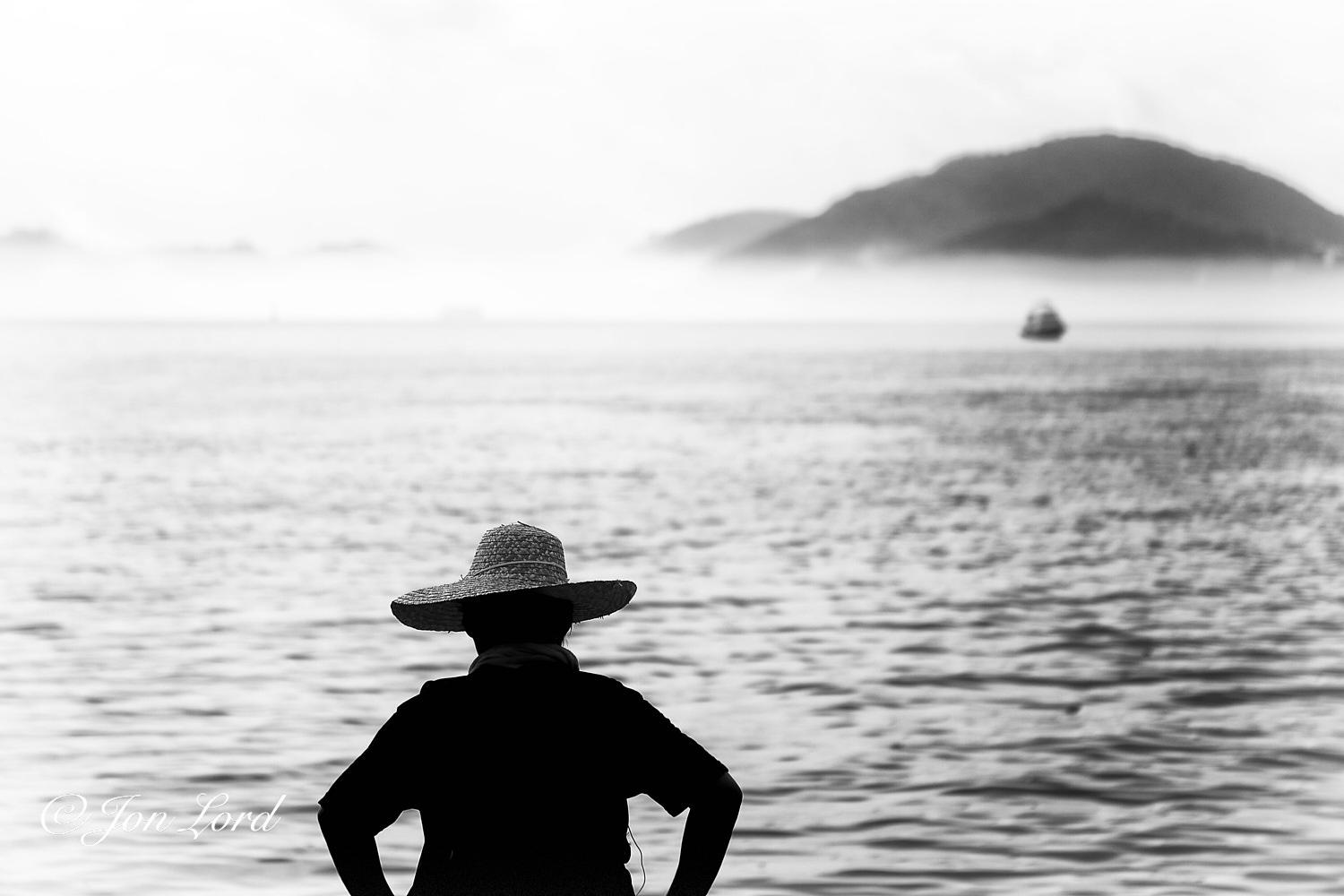 This is a black and white street photo in landscape format of the silhouette of a person who is looking out to sea with a bank of sea fog in the distance and an island rising out of the fog. Mui Wo, Hong Kong (2013).

The rippled, silver and black sea water rises from the base of the photo upwards to the three-quarters up level. Slightly to the left and below centre is the silhouette of our person. They are viewed from behind at a distance of a few metres with them filling a little over one third of the height of the image. They are standing at the waters edge on a small harbour wall that is out of view below. The person is viewed from the waist up, is wearing a dark short sleeved shirt and is all in deep shadow. The only item not in shadow is a wide brimmed straw hat that is very slightly tilted to the left. Beyond our person is an expanse of sea with a single small boat above and to the right of centre. Behind the boat is that bank of sea fog that rises perhaps only ten metres above 
