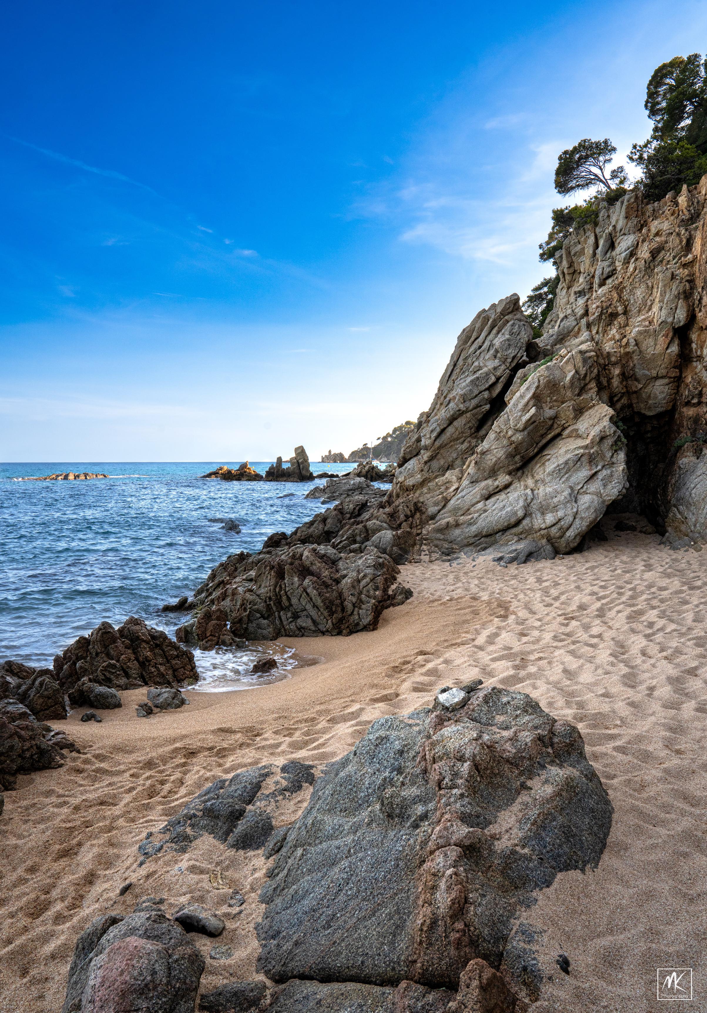 Color photo of a rocky beach on a blue sea under blue skies with cliffs in the background and rocks out in the water.