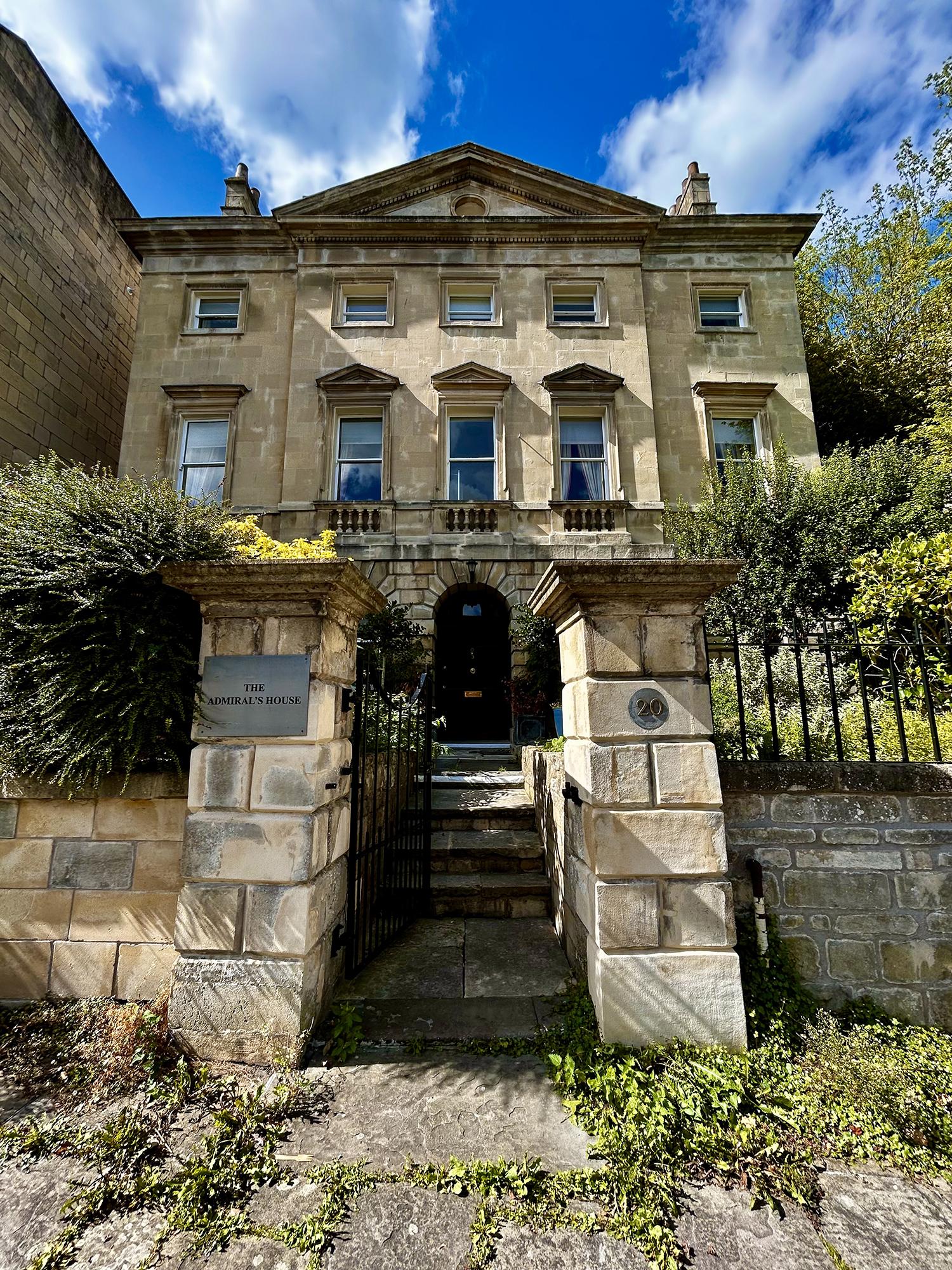 A large, detached Georgian house and its gateposts on a summer's day. This three-storey, five-bay fronted Palladian villa is topped by a large pediment. Its six-panel door has a fanlight and the first floor has balustraded panels below the windows. The front garden has lush trees and shrubs in various shades of green. In the foreground, outside the property, an abundance of weeds grows between paving slabs. A modern sign on the gatepost reads "The Admiral's House" and you can rent the seven-bedroom property via a popular holiday accommodation website. 20 Vineyards (c. 1765), The Paragon, Bath, UK. Photo by Gearóid Burke.