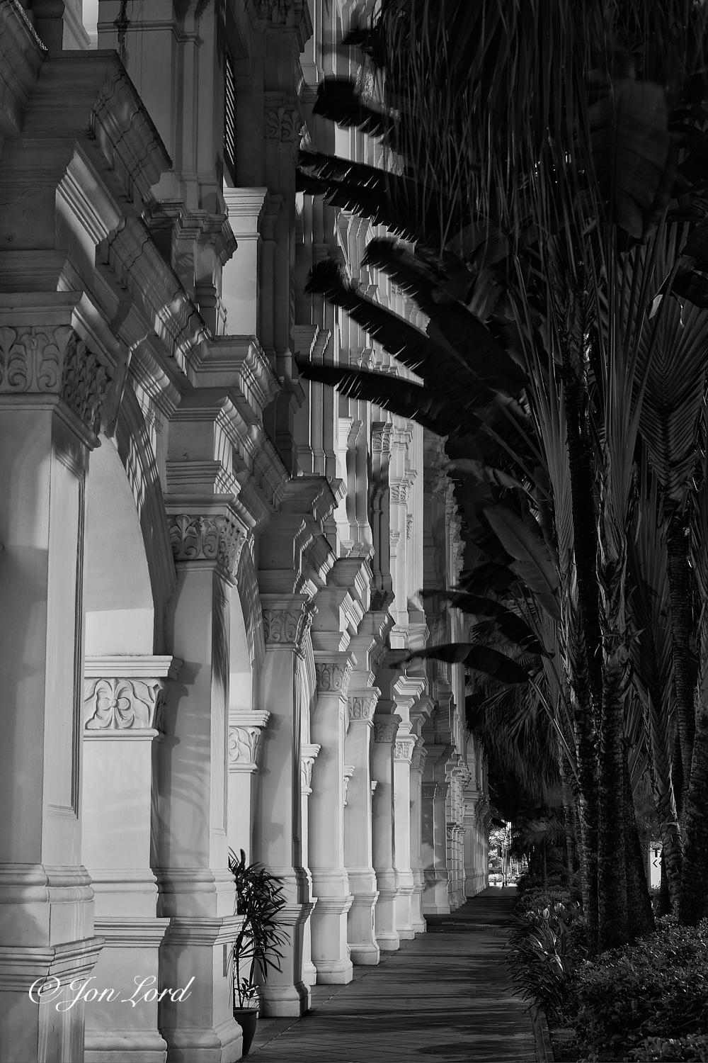 This is a night time photo in portrait format looking along a deserted pavement with an ornate white facade lined with pillars on the left side and a row of palm like trees on the right. North Bridge Road, Singapore (2013).

Extending away from the camera from the centre of the base of the photo into to distance and angled to the right is a dark, two metre wide pavement. Commencing along the left edge of the image and stretching away from the camera and again, to the right is the white facade of a restored historic building. The camera is placed two metres away from the front of the building and is angled along the facade so that the gaps between the pillars are visible but nothing can be seen inside those gaps. The facade is lined with ornate white and decorated pillars placed every few metres, the arched gap below allowing entry into the building, it's courtyards or the shops that are on the outside of the building. Above the pillars and arches is a tiered plinth that stretches the l