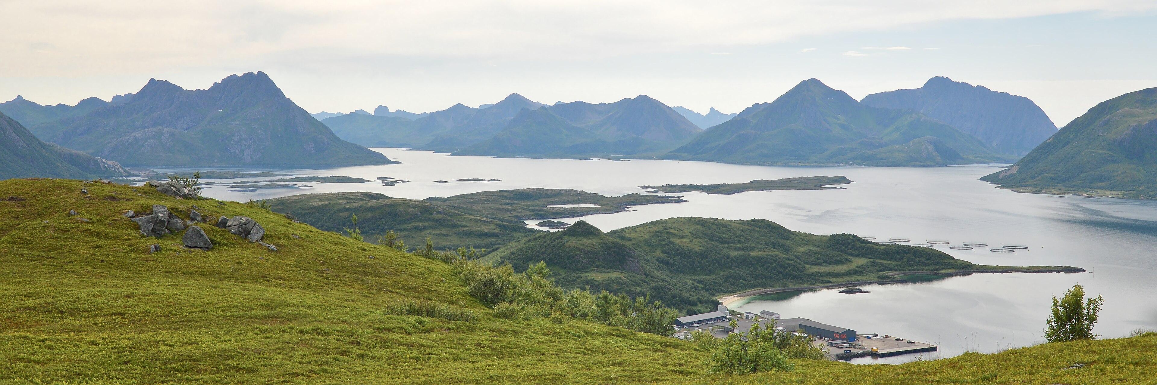 A panoramic photo looking off the side of a mountain at a peninsula with mountains in the distance. The sky is filled with clouds.