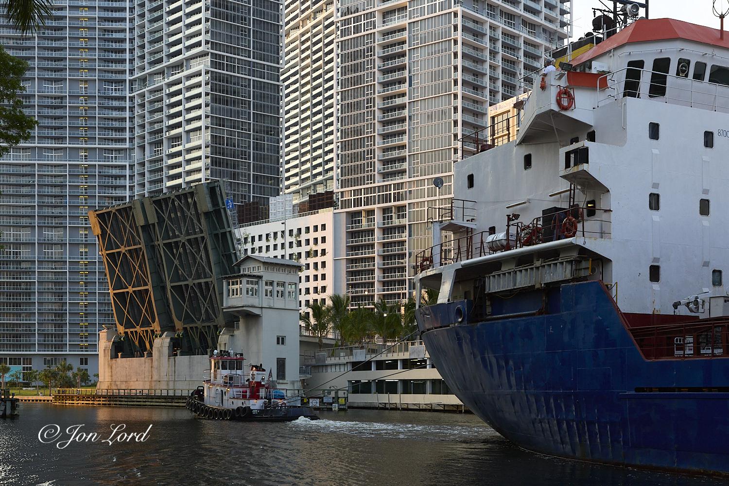The is a dawn colour photo in landscape format of a small coastal cargo ship being towed backwards along a city river surrounded by high-rise apartment buildings with the raised Brickell Avenue bascule road bridge ahead. Miami River, Florida (2012).

The lower 10% of the image is filled with dark and slightly rippled river water. Extending from the right edge of the photo to the centre is the aft section of the small to medium sized cargo ship, with the stern portion of the medium blue hull and white superstructure in view. The ship fills the entire height of the image, half the width and is viewed from 10m away. The blue hull, while clean is very dented with numerous ribs visible around the depressions in the hull. Above the rounded stern is the accommodation and wheelhouse superstructure with 3 decks of small square windows below the wheelhouse. To the left and below centre is a tug underway from right to left and attached to the ship with 2 stout lines. Behind the tug is a long whit