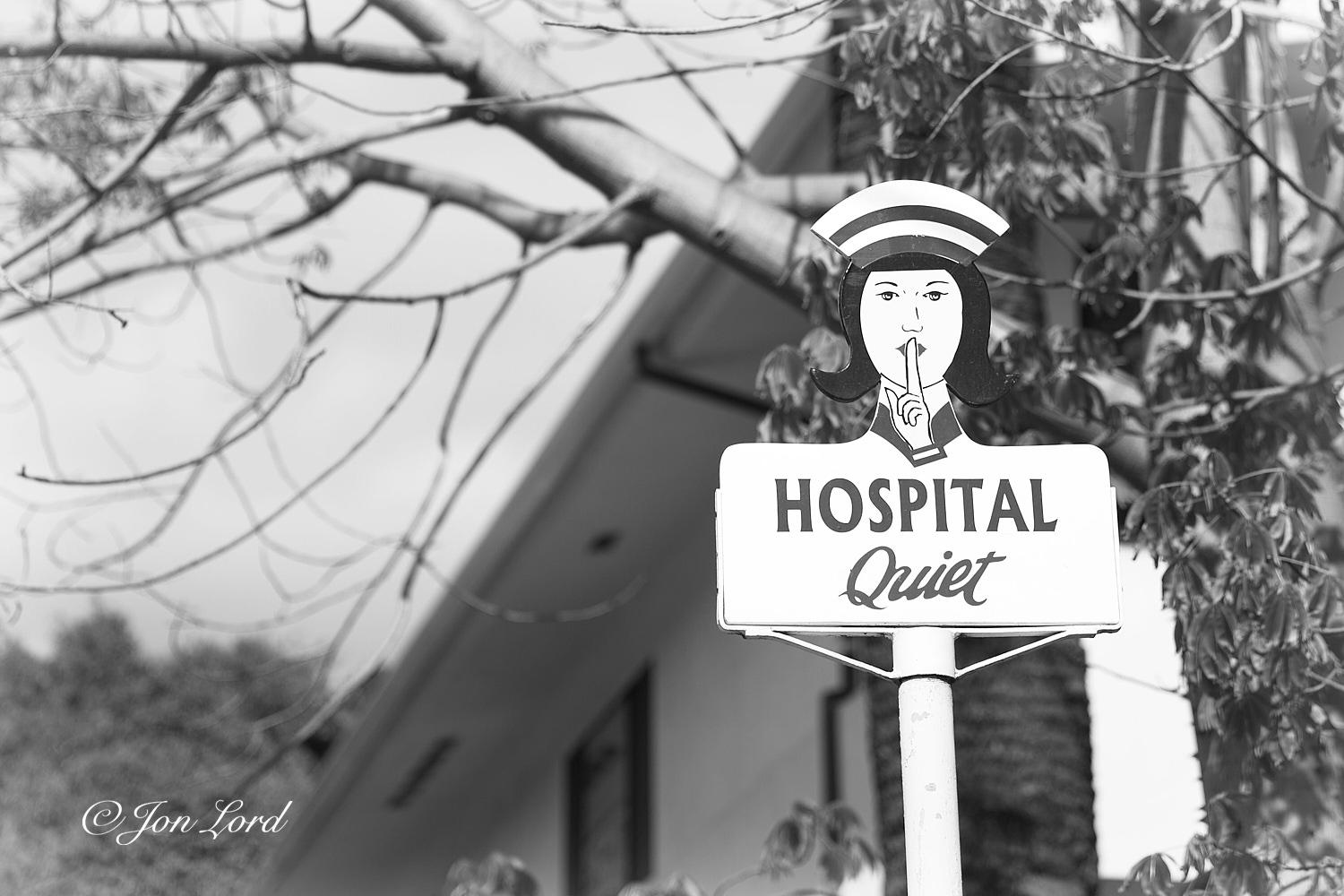 This is a black and white rural street photo in landscape format of a road sign with the cutout of a 1950's style nurse with her hand raised to her face and a finger over her lips with the words below in large letters 'HOSPITAL'  and below in smaller Italic lettering 'Quiet'. Avalon, Santa Catalina (2014).

To the right of centre and mounted on a long metal pole is our sign. The camera is angled upwards and to one side of the sign and at a distance of about two metres with the sign filling three-quarters of the height of the photo. On top of the metal pole is the wooden cut out of the nurse and the rectangular white sign with the lettering in black below the nurse. The centre of the upper part of the sign rises upwards to form the nurse's neck with a black collar, serious looking face above, dark hair curling around the ears and an old fashioned nurse's hat or cap. Her left hand is held up to her face with her index finger raised up to her pursed lips. Behind the nurse is a tree growin