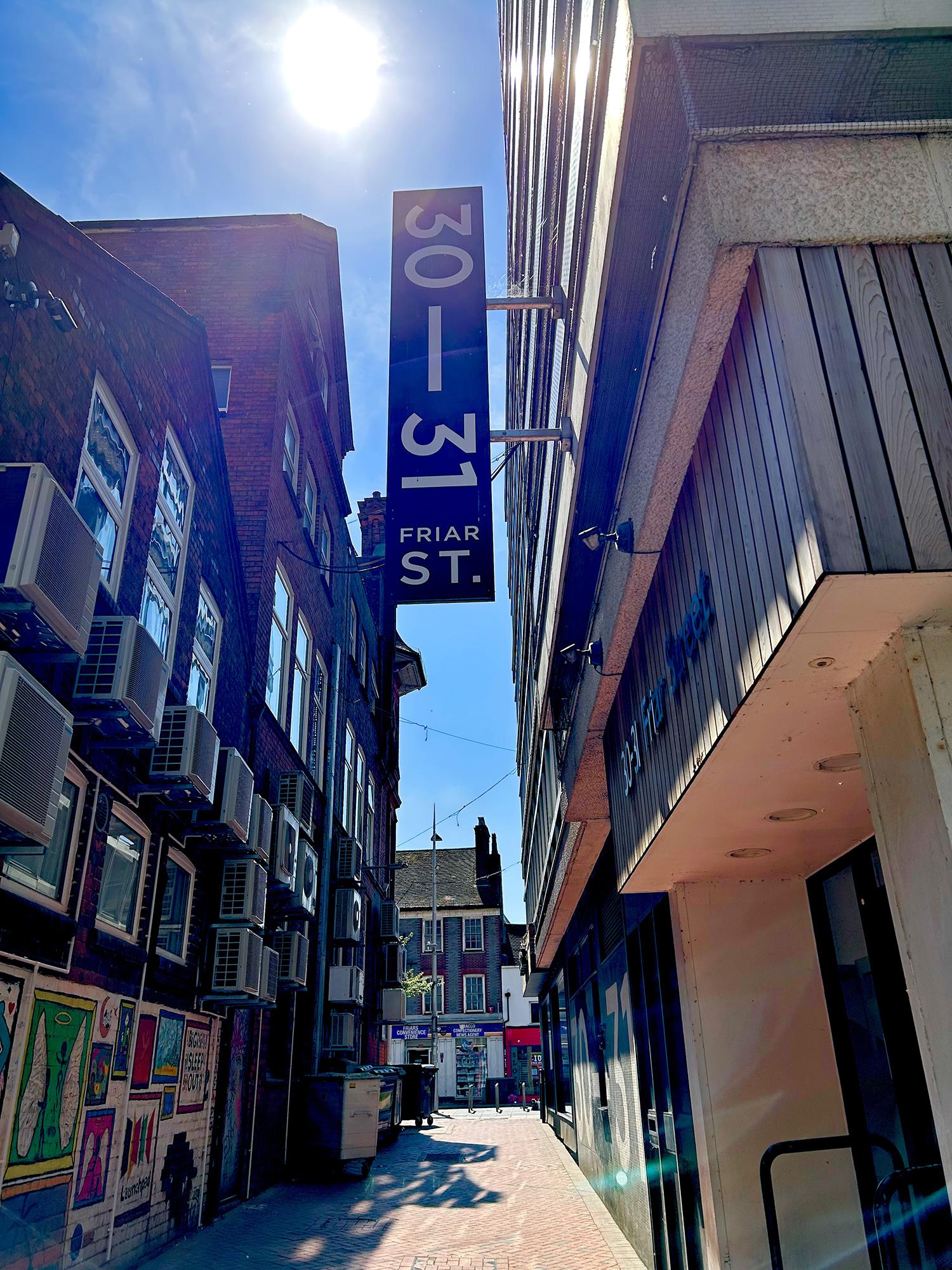 An urban passageway. The old brick buildings on the left feature many white-framed windows and numerous air-conditioning (or perhaps heat pump) units. Refuse bins are lined up along side it. On the right, a relatively modern office/retail building, with concrete walls, wood panelling, and a mesh-screened balcony. Attached to the latter is a vertical hanging sign that reads "30—31 Friar Street." The em-dash is stylistically wrong (it should be an en-dash), but I don't mind it. Reading, Berkshire, UK. Photo by Gearóid Burke.