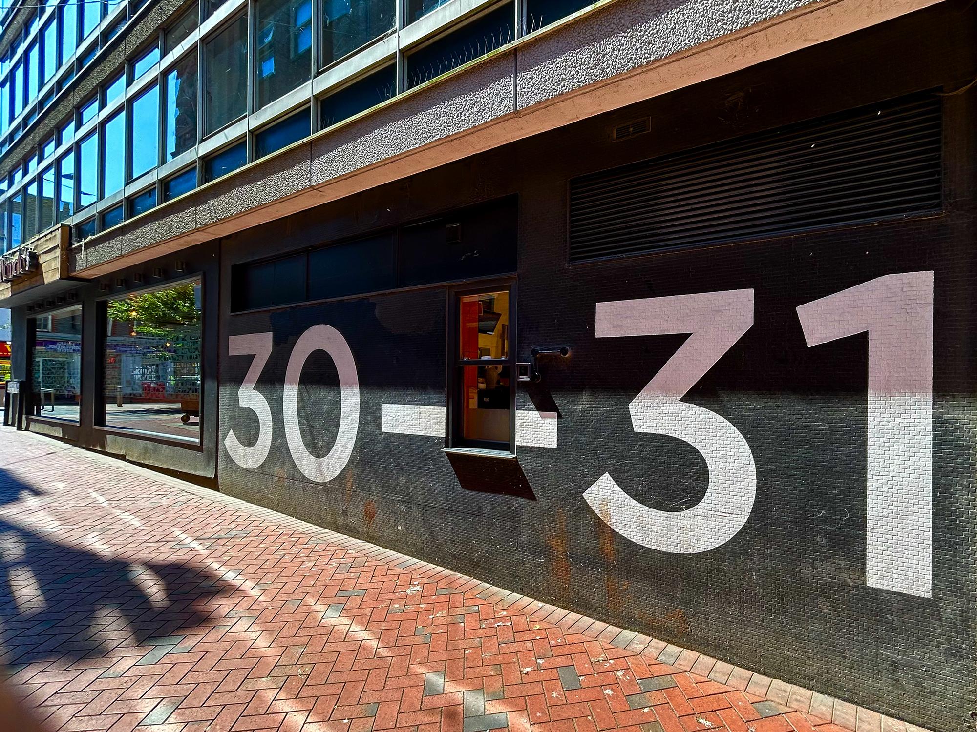 In a paved alley, one section of an otherwise concrete-and-glass building has a brickwork section with a small sash window in the middle. The wall has been painted black and has "30—31" painted in large white numerals, with the extra-long dash being broken by the window. 30–31 Friar Street, Reading, Berkshire, UK. Photo by Gearóid Burke.