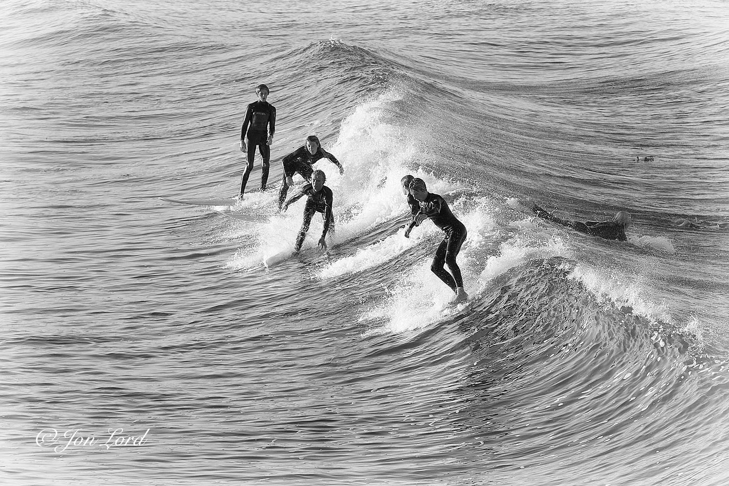 This is a black and white photo in landscape format of a group of young teens surfing shortly after sunrise. Hermosa Beach, California (2013).

Descending from the upper left corner diagonally across the the photo down towards the lower right corner is a wave that rises upwards about two metres and starts to curl from the centre of the photo downwards and is in motion from right to left. Scattered around the centre of the image is the group of five young male teens, all dressed in black wetsuits and standing on their surfboards ahead of the breaking wave. The young surfers are viewed from above and at a distance of ten to fifteen metres. The surfers are in a line across, each separated by a metre or two. The closest teen to the camera is crouched very slightly as he surfs across the wave and towards the camera. The second is partially obscured by the first and appears to be surfing across the wave and away from the camera. The third is fully crouched down, arms spread out to maintain b