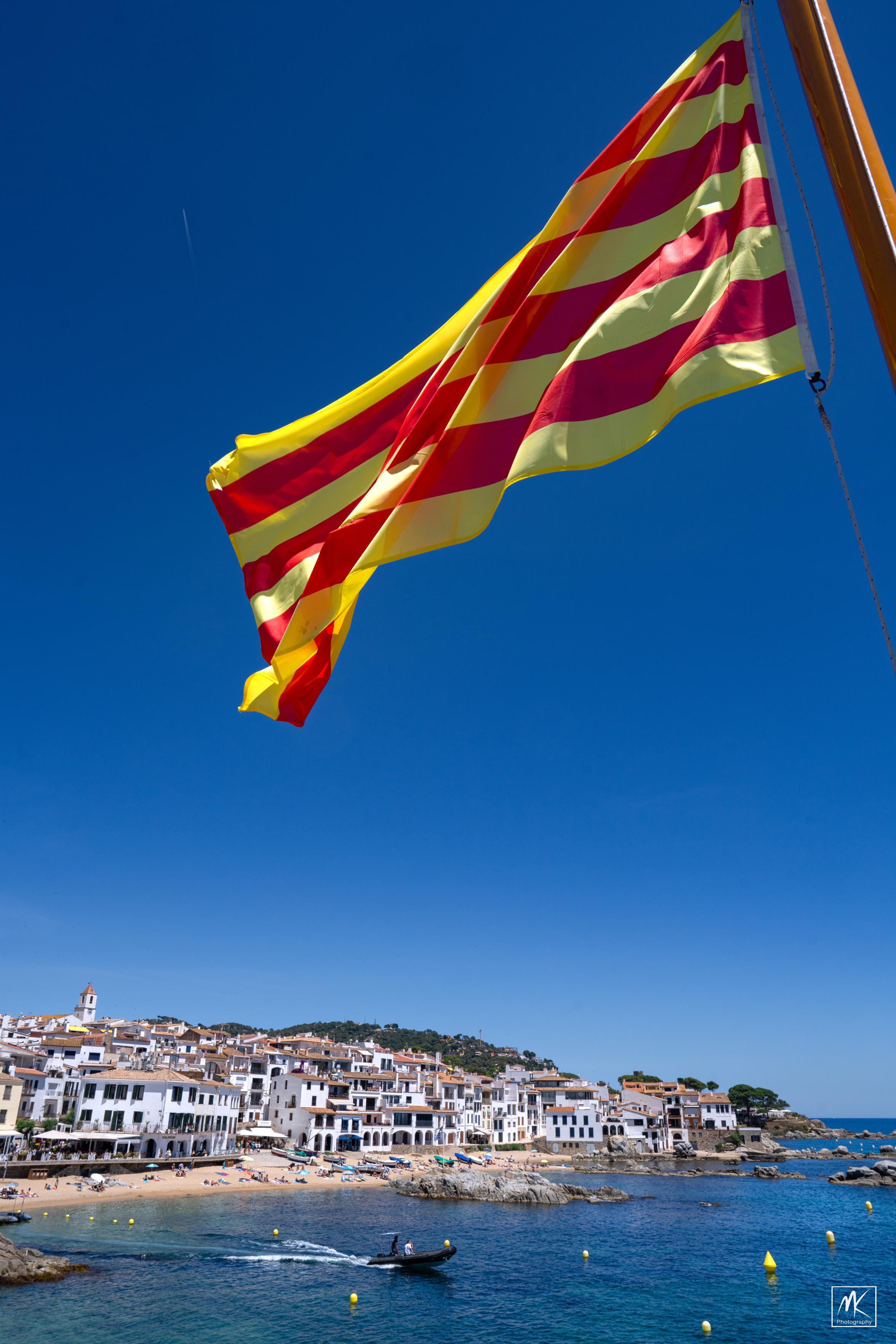 Color photo of a seaside town of white buildings under a blue sky with a sandy and rocky beach in front of it. There is a pontoon boat heading out from the shore and a large Catalonian flag with red and yellow horizontal stripes hanging from a pole at the top of the frame in the foreground.