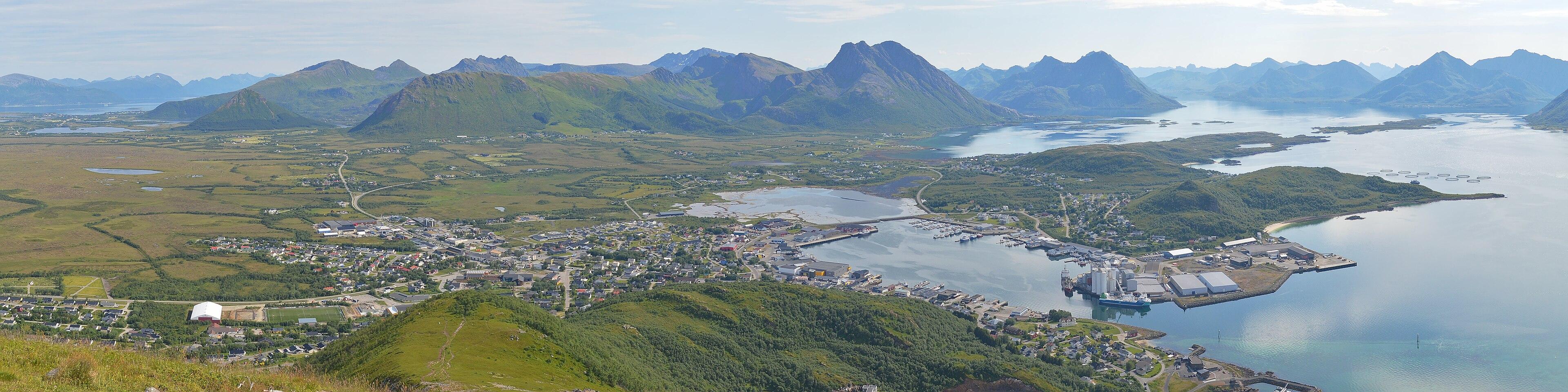 A panoramic photo looking down from a mountain at a town. There is a peninsula to the right and wetlands to the left. There are mountains in the distance. The light is hazy and the sky has clouds in it.