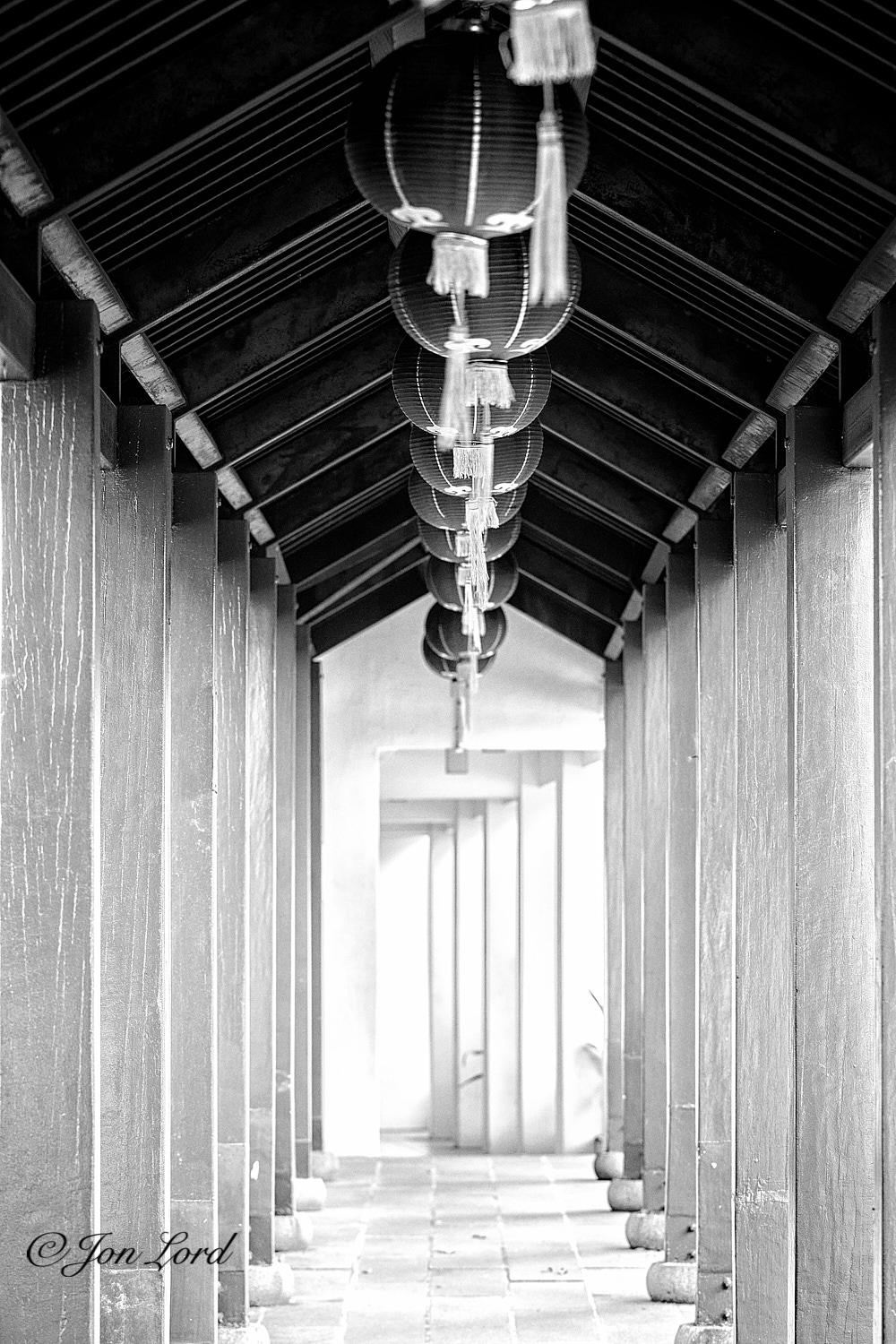 This is a black and white: part abstract, part historic, part architectural photo in portrait format of a narrow covered walkway with square wooden pillars supporting a pitched roof with multiple Chinese lanterns suspended from the centre of the inside of the roof. Old Thong Chai Medical Institution, Singapore (2013).

Extending away from the camera along both the left and right sides of the photo are two rows of square, tall, wooden pillars that are about two and a half metres tall and with the two rows spaced about by one and a half metres. The pillars converge slightly with increasing distance. The wood is a pale white in appearance, perhaps bleached, painted white or the elapse of time? At the base of the pillars are round stones with a flat top and bottom, identical to a wheel of cheese. The floor comprises of multiple paving stones that are not quite laid flat, again perhaps suffering the passage of time. On the top of the pillars is the inside of a narrow pitched roof. The roof 