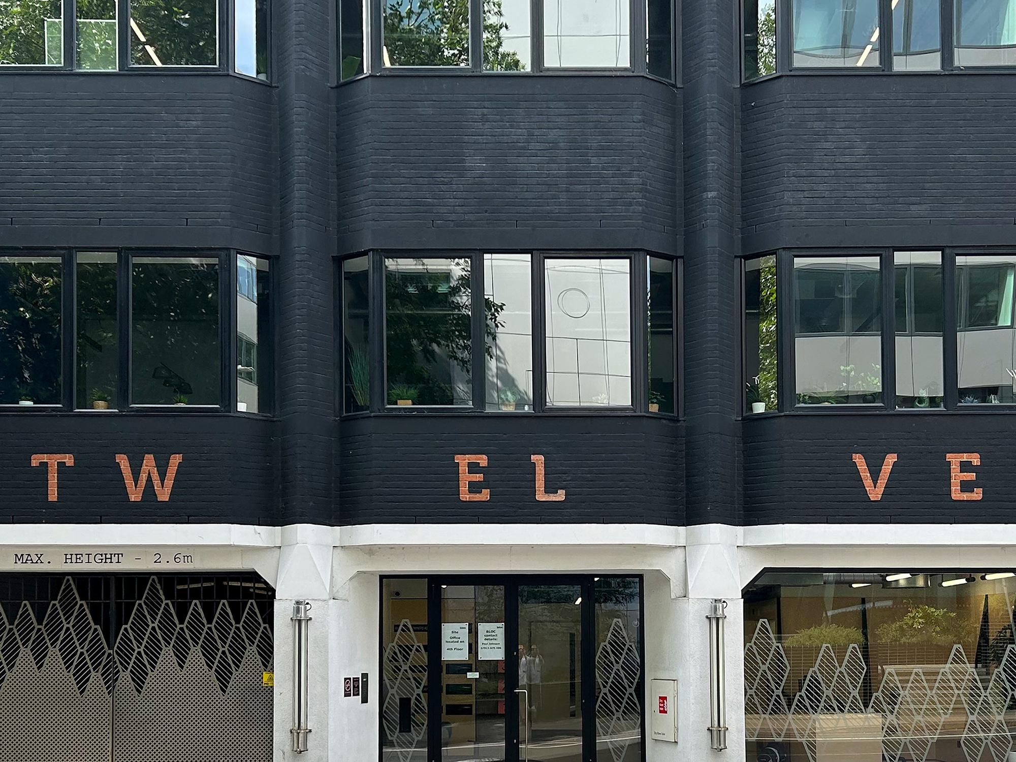 A redbrick office building has been painted black, except for its stylised street number ("TW EL VE"). On the street level to the left, the metallic gates to the structure's parking garage feature an interesting geometric pattern which is mirrored by a screen behind the reception area window on the right. 12 Forbury Road, Reading, Berkshire, UK. Photo by Gearóid Burke.