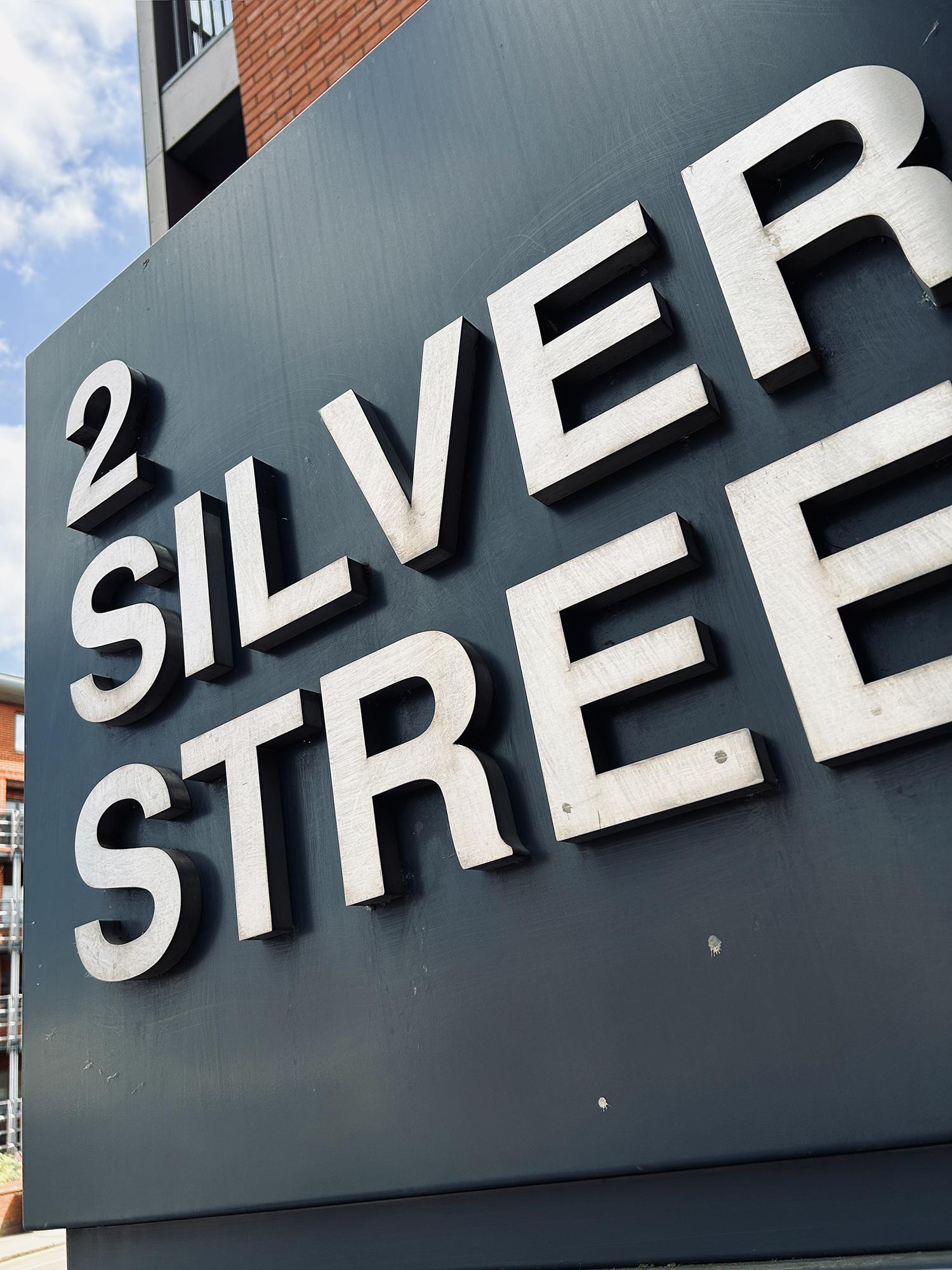 2 Silver Street: the freestanding, monolith-like sign for this apartment building is made with dark grey metal panels and has protruding stainless-steel lettering (plus one numeral). Taken at an angle, this photo crops out the final letter T. The metal has weathered a little and the letters are somewhat discoloured. A blue sky with wispy clouds is visible in the top left corner. Reading, Berkshire, UK. Photo by Gearóid Burke.