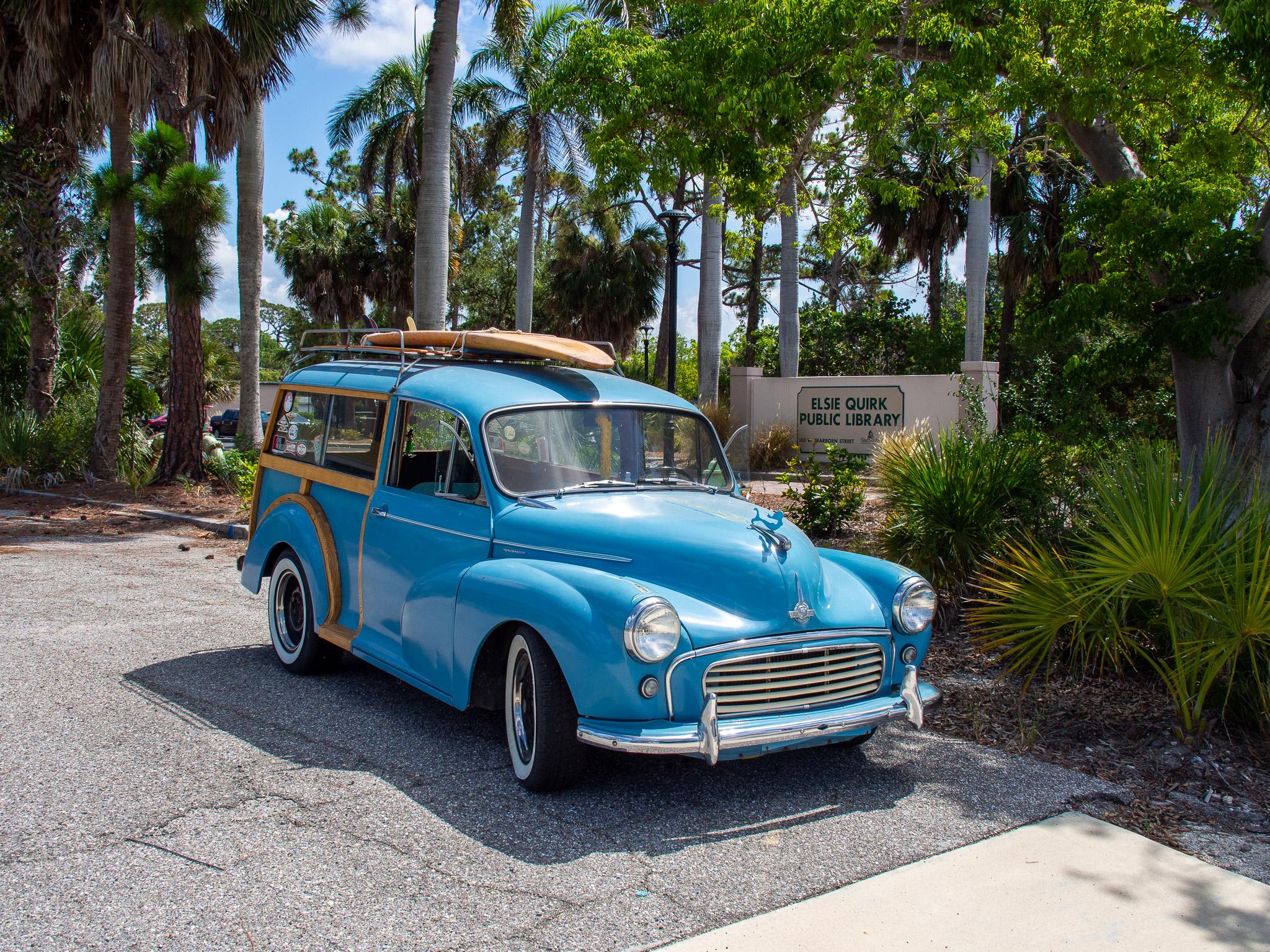 Old blue van with round headlights, curved hood, and curved fenders. There is a lot of chrome trim, and wood trim around the rear of the vehicle. There is a mermaid-shaped hood ornament, and a wooden surfboard on a rack on the top. The tires are whitewalls. It is sitting on asphalt pavement. In the background are palm trees and other trees, and there is a sign behind the car that says "Elsie Quirk Public Library". It is a sunny day and there is some blue sky with clouds in the background.