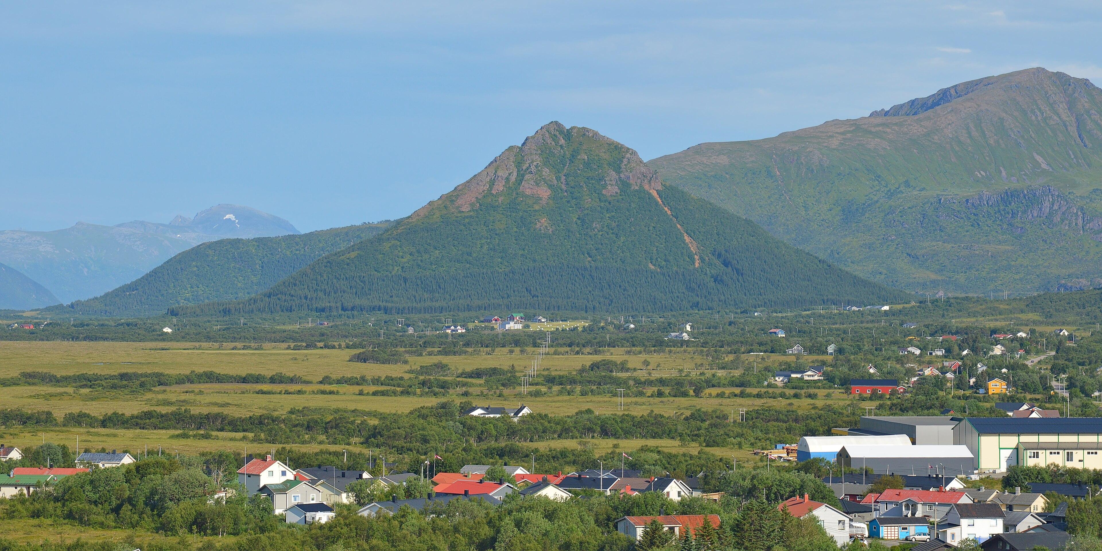 A photo of a pyramid shaped mountain with more mountains behind it. The sky is blue.
