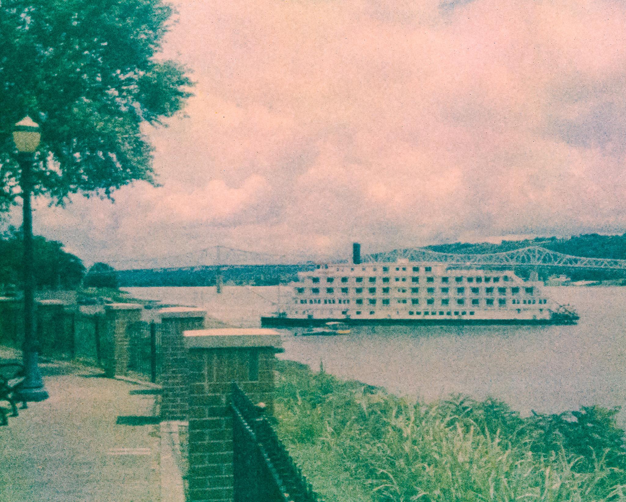 On the left in the foreground, a walkway and a fence made from brick and iron. Also an old-style streetlamp, and some trees. In the foreground on the right a grassy, sloping riverbank. Near the center of the frame a large river with a large riverboat with many windows/doorways, a smokestack, and a paddle wheel at the back. There is a smaller boat in front of the larger one. In the background a long bridge crossing the river, and some distant trees on the far side of the river. The sky is cloudy. The photo is very grainy, and the colors are muted. The sky has a pinkish tint.