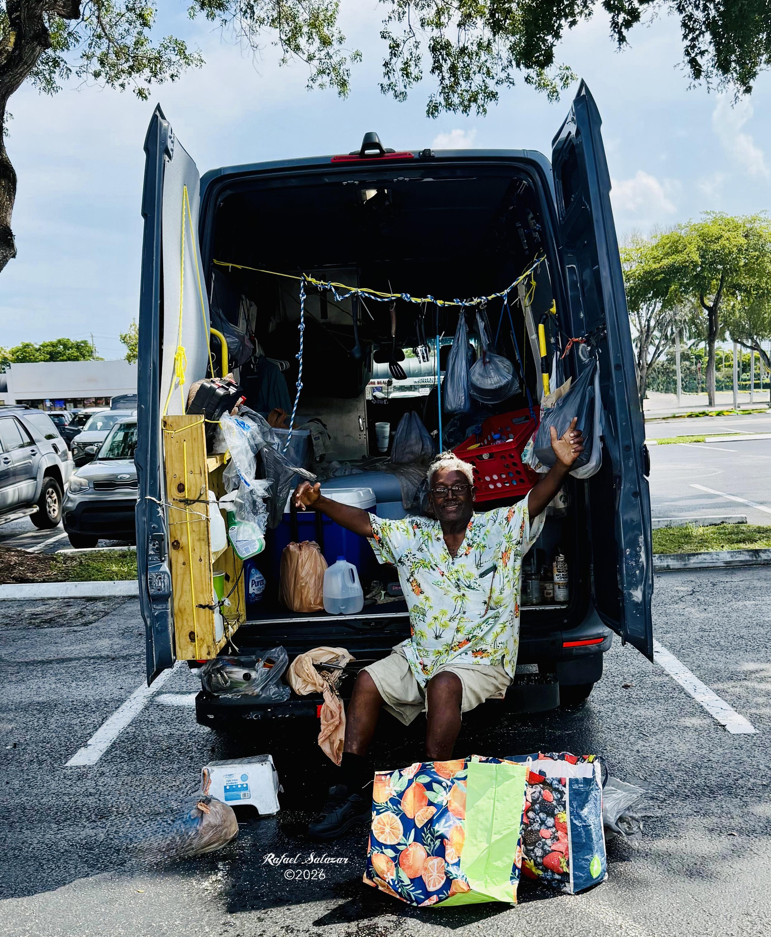 Documentary photograph by Rafael Salazar showing a man seated at the open rear of a blue Mercedes-Benz Sprinter van in a Florida parking lot, surrounded by bags, containers and personal belongings, raising his arms toward the camera beneath a bright summer sky.