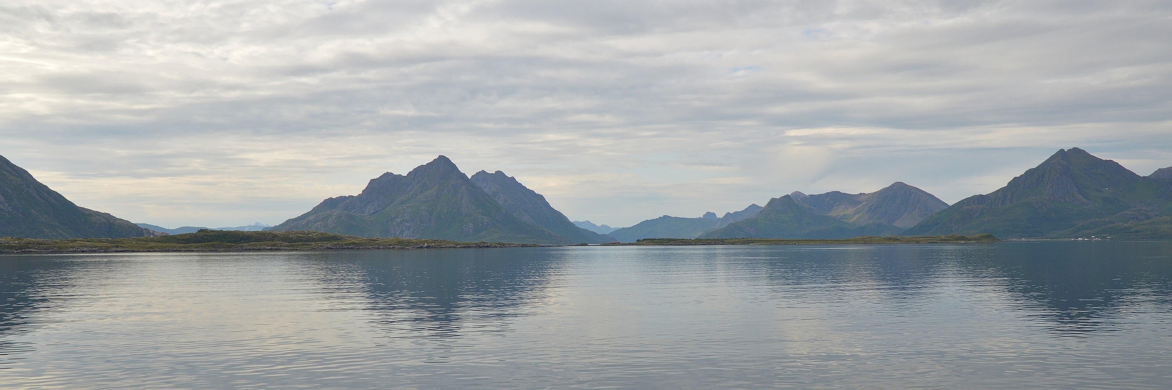 A panoramic photo of water with mountainous islands in the distance. The sky is filled with clouds.