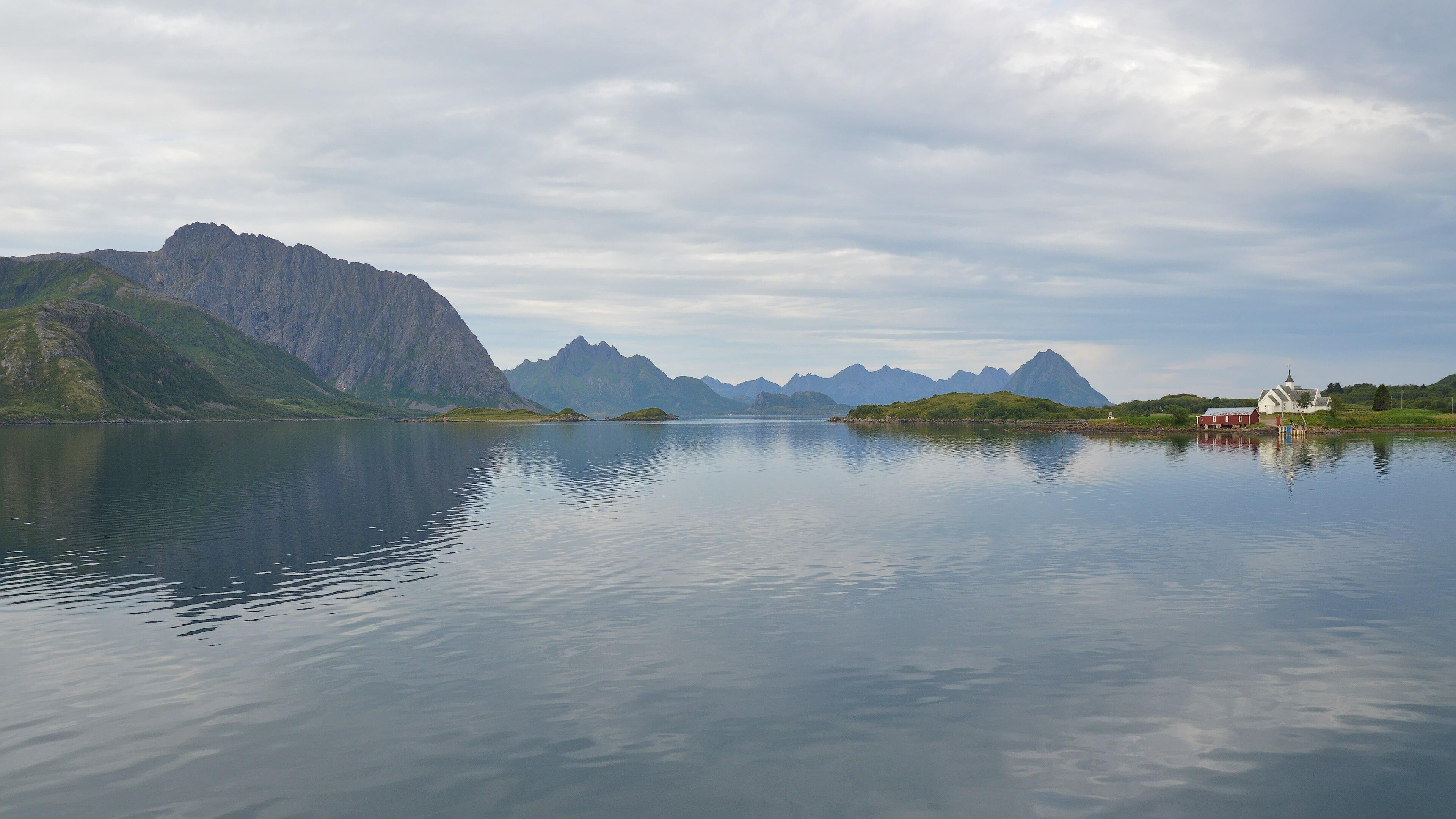A photo of a strait with mountainous islands on the left. There are a red building and a white building on the shore on the right. The sky is filled with clouds.