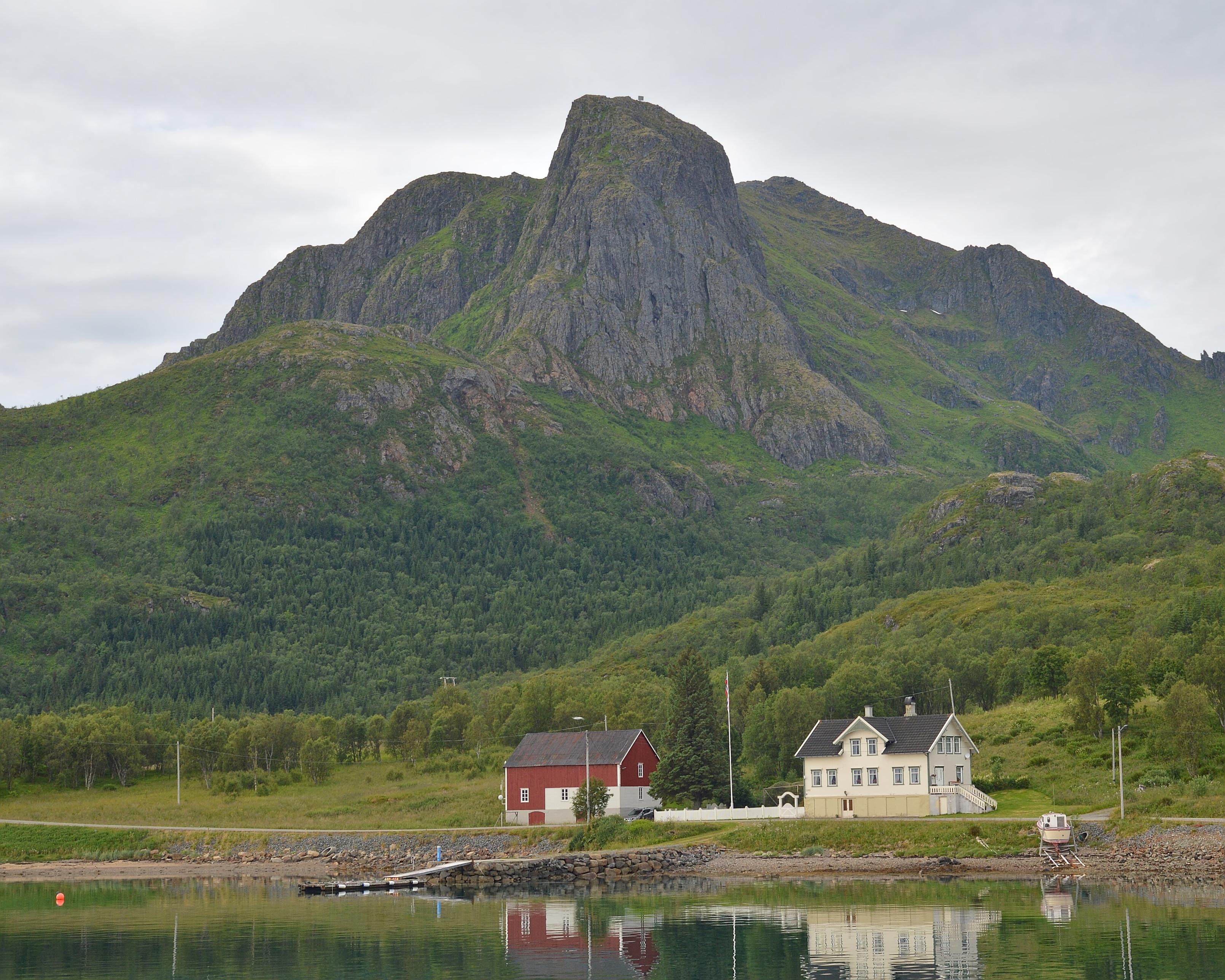 A photo of a mountain with a red building and a white building on the shore below it. The sky is filled with clouds.