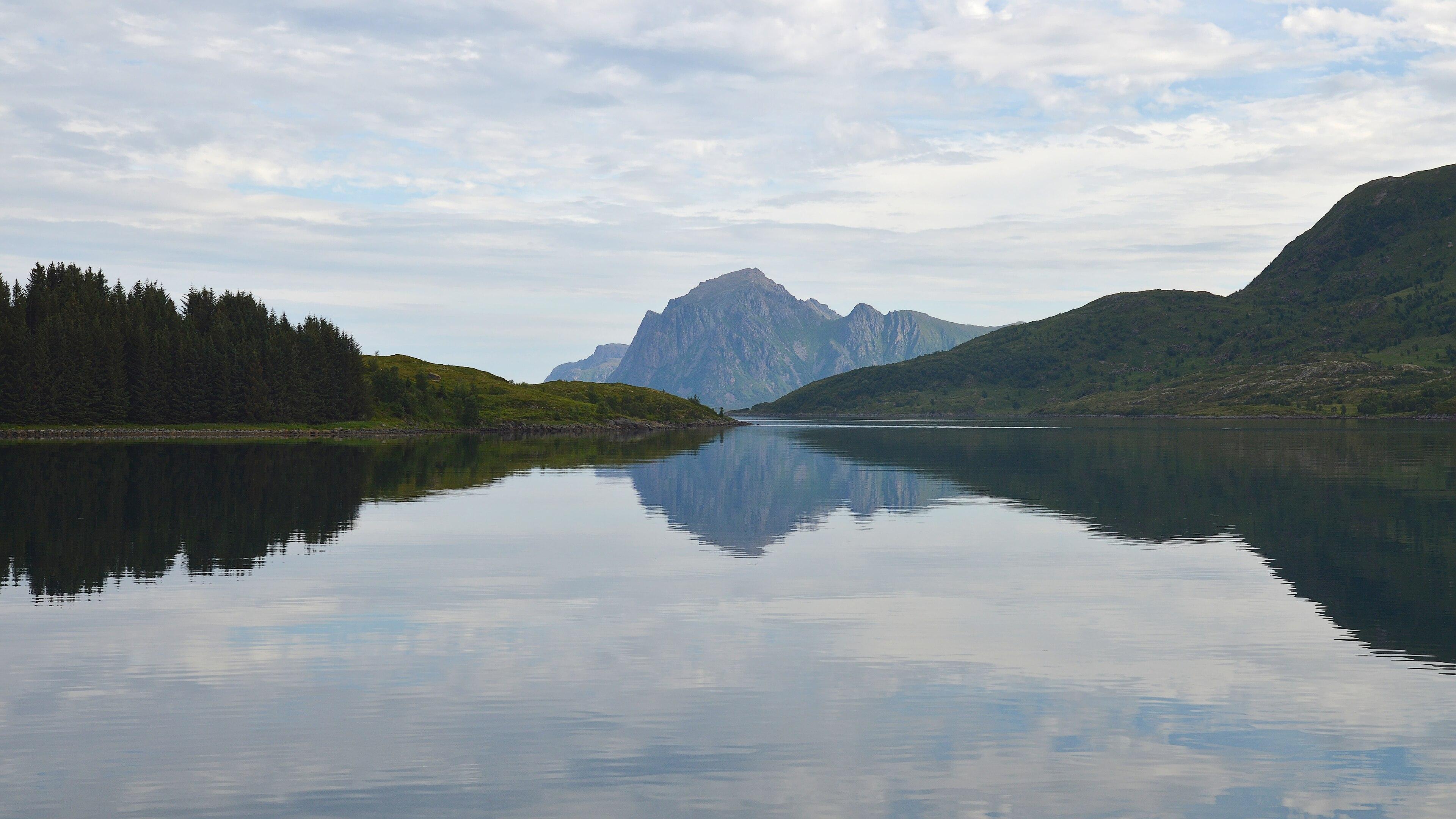 A photo of a strait with the ends of two islands lined up in the middle. There's a mountain in the center behind them. The water is still and the sky is filled with clouds.