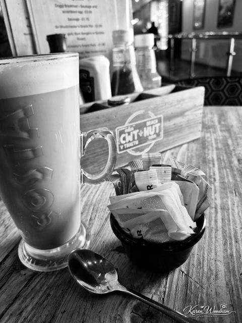 Cafe table setup with a tall latte in a glass mug, a small bowl of sugar packets and a spoon on a weathered wooden tabletop, condiments in a wooden caddy blurred in the background.