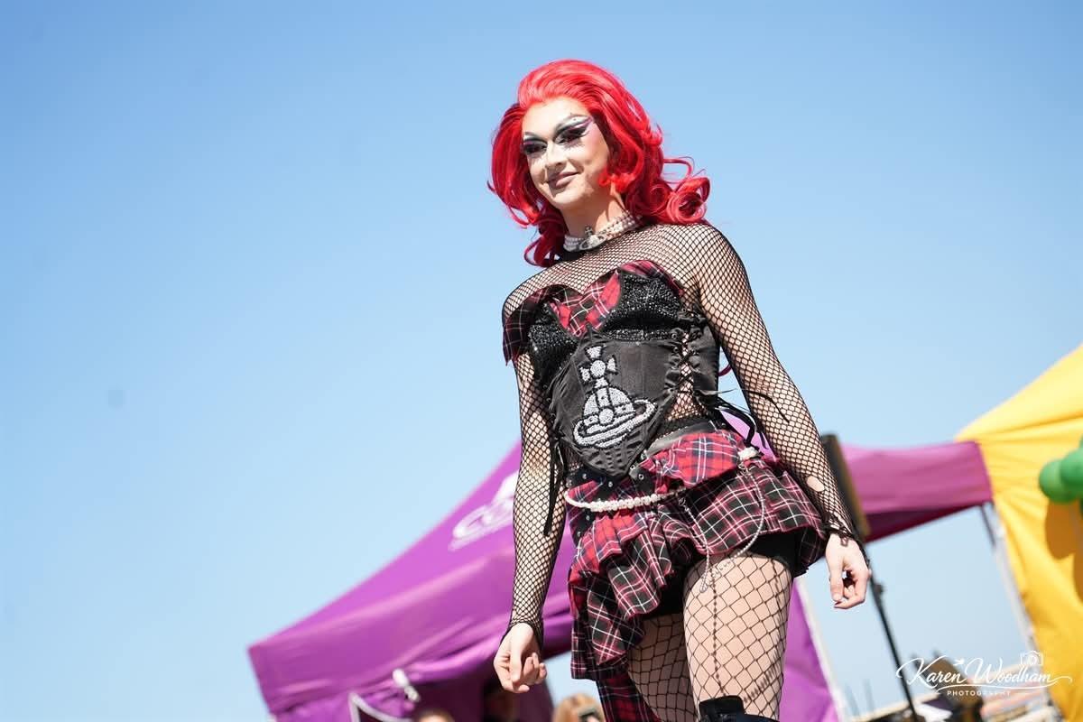 Drag performer in a red wig and dramatic makeup wearing a black corset, plaid skirt and fishnet sleeves and stockings, smiling confidently while posing on an outdoor festival stage under a clear blue sky.