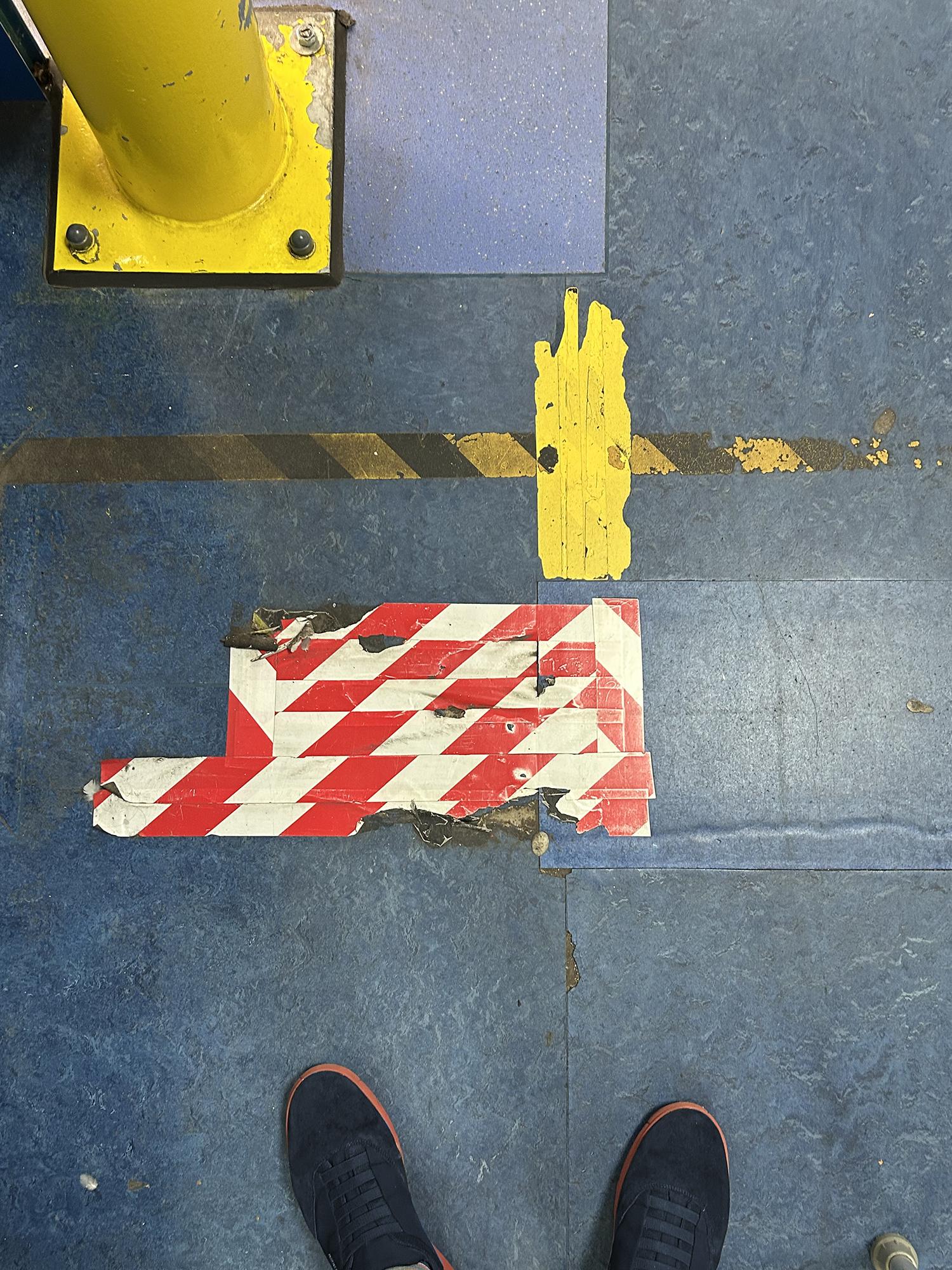 An aged, blue linoleum floor features numerous strips of hazard tape and paint. The central grouping of these adhesive applications has six horizontal lines and two vertical lines of red-and-white striped tape. There's a bright yellow bollard in the top left corner, and two shoes with blue uppers and red soles at the base of the image. Royal Berkshire Hospital, Reading, UK. Photo by Gearóid Burke.