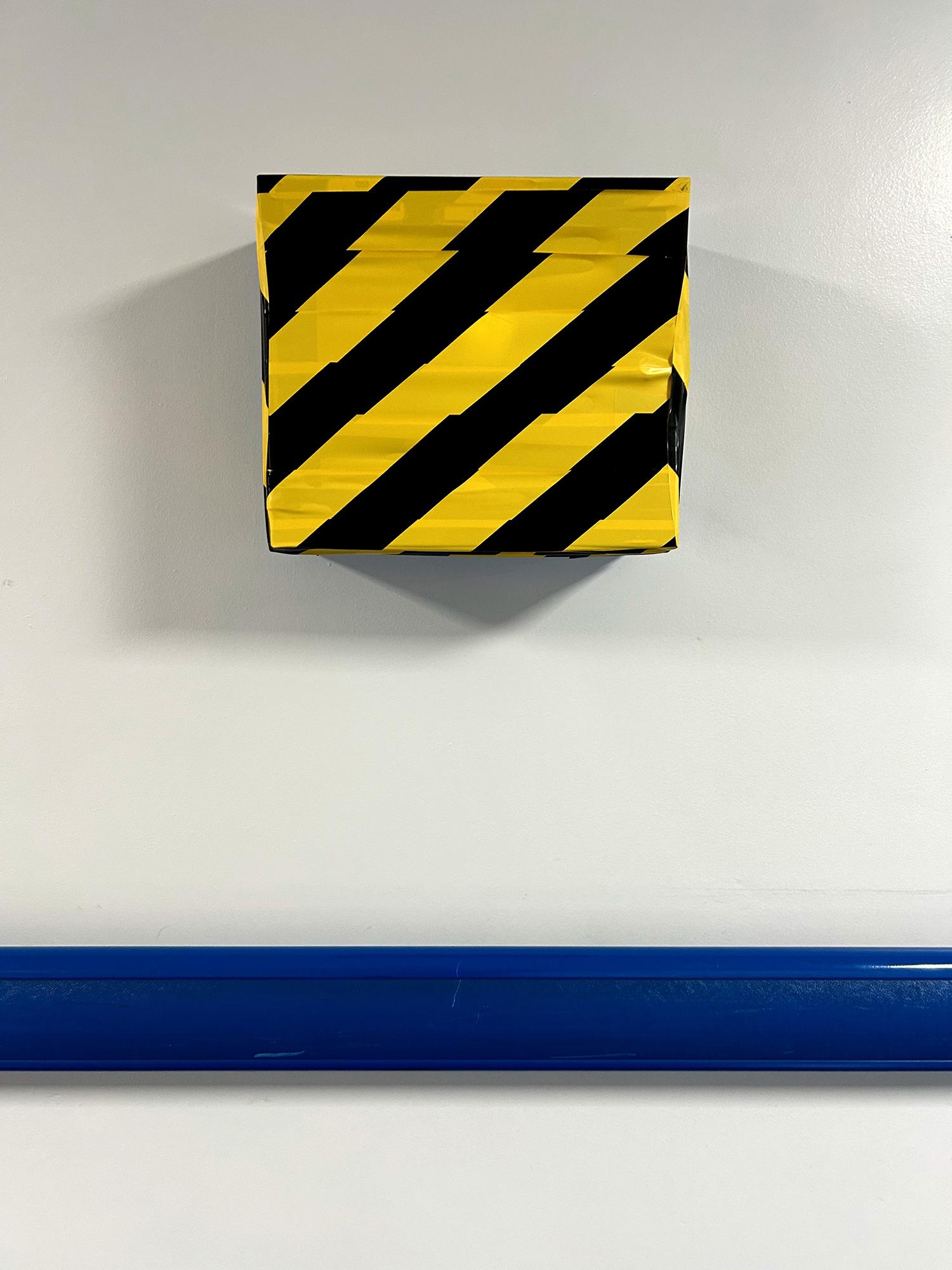A wall-mounted box has been completely covered in yellow-and-black hazard tape. The wall itself is painted white and ceiling lights cast shadows to the left and right. A blue plastic handrail is below the box. United Kingdom health and safety regulations state that markings should be placed around obstacles or dangerous locations. This includes where any of the following present a risk: people tripping or falling; objects falling, and people or vehicles colliding with objects. Royal Berkshire Hospital, Reading, UK. Photo by Gearóid Burke.
