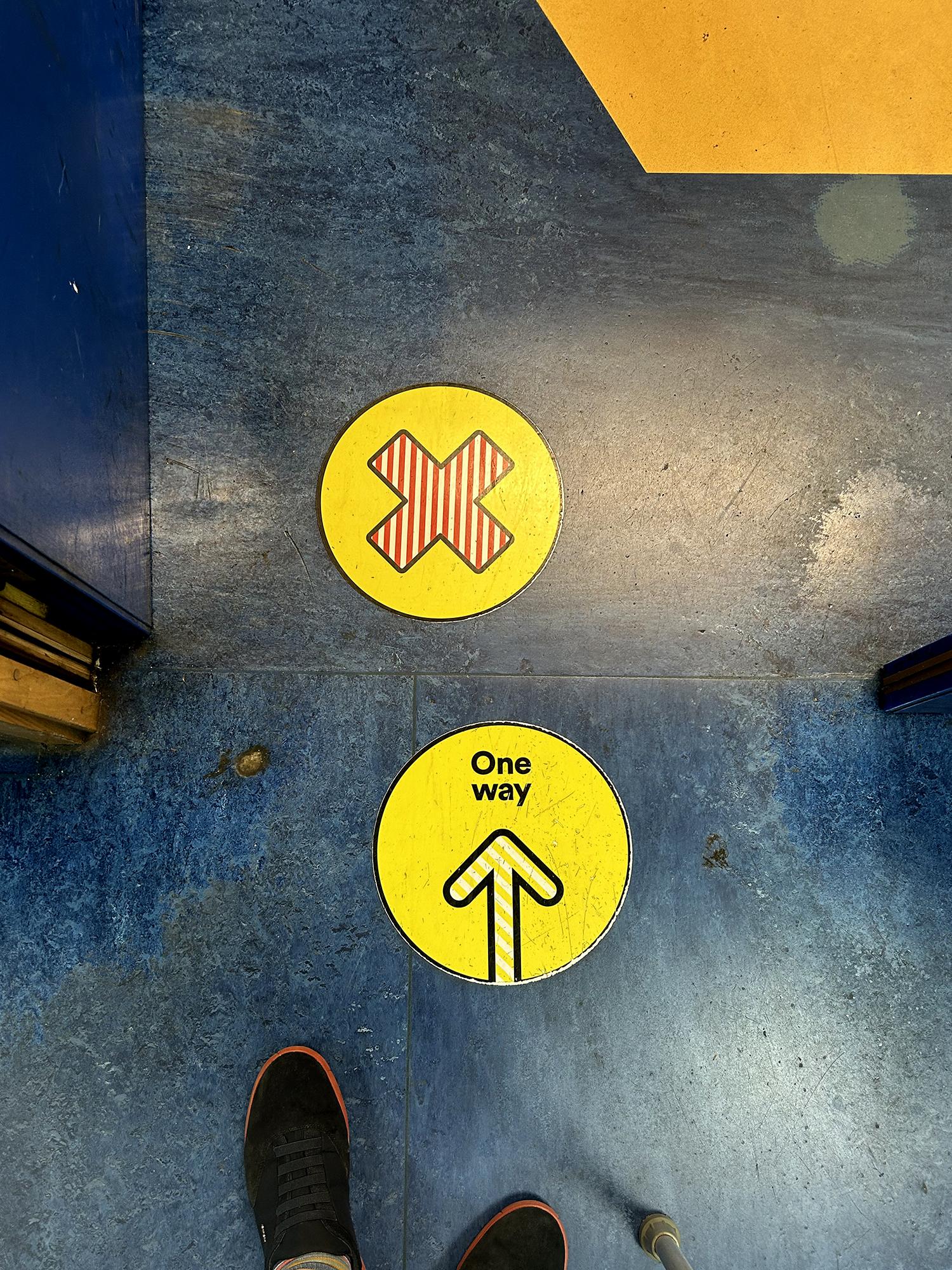 Standing at a doorway and looking down to aged and stained blue linoleum. There are two stickers on the ground, both mostly yellow in colour. One has an x, or more accurately, a multiplication sign (×), filled with a red and white stripe pattern. The other reads "One way"  and has an upward point arrow filled with white and yellow stripes.  There's a polygon of warm yellow linoleum in the top right corner, and two shoes with blue uppers and red soles at the base of the image. Royal Berkshire Hospital, Reading, UK. Photo by Gearóid Burke.