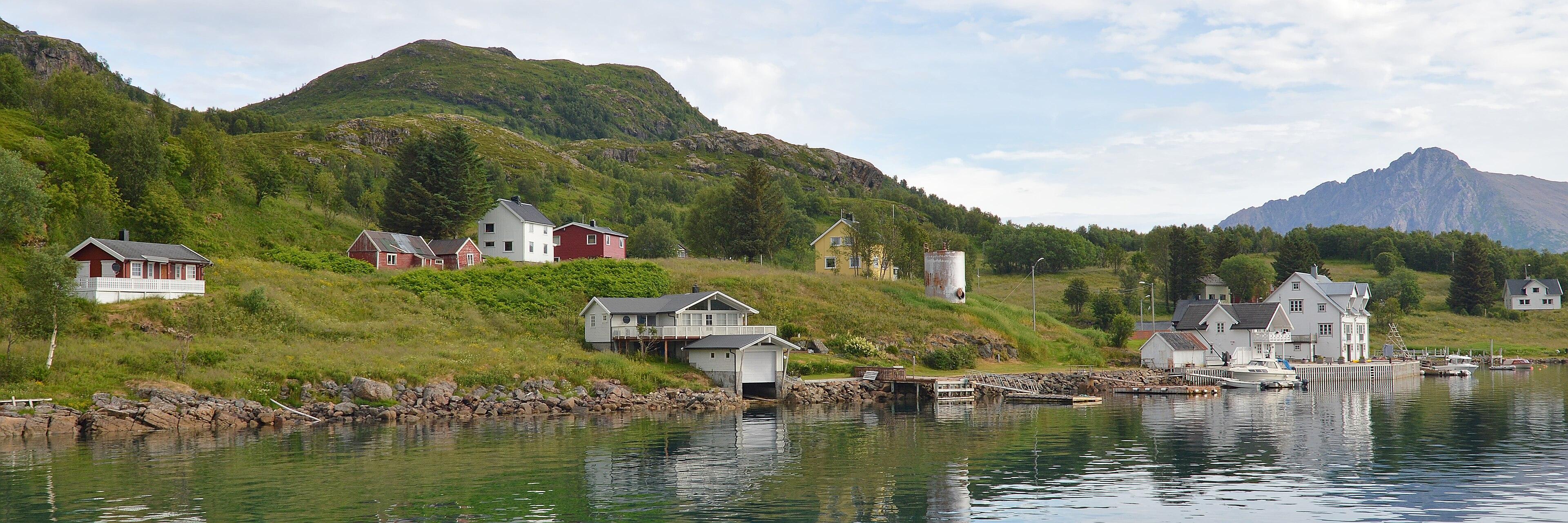 A panoramic photo of a small village on a shore with mountains behind it. The sky is blue with white clouds in it.