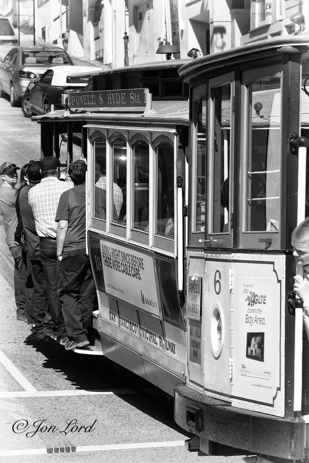 This is a black and white photo in portrait format of a San Francisco Cable Car cross roads with one cable car moving a right angles across the camera and the other travelling away. San Francisco, California (2013).

Extending from the right edge of the photo to the centre is the front of the first cable car, viewed from no more than two metres away. Peering around the front is the head of a young boy, holding onto a white vertical hand hold. The base (chassis) is black metal with the wheels out of view below the image. Above, on the front, are three rectangular wooden panel. The near panel has a small advert. The centre has a single headlight and the number 6 above while the far panel has the route of the cable car. Above are three vertical rectangular windows with the roof above. On the far side of the cable car is the other further identical cable car, in motion away from the camera and at right angles to the first. The left side is in view with the rear obscured by the first car. I