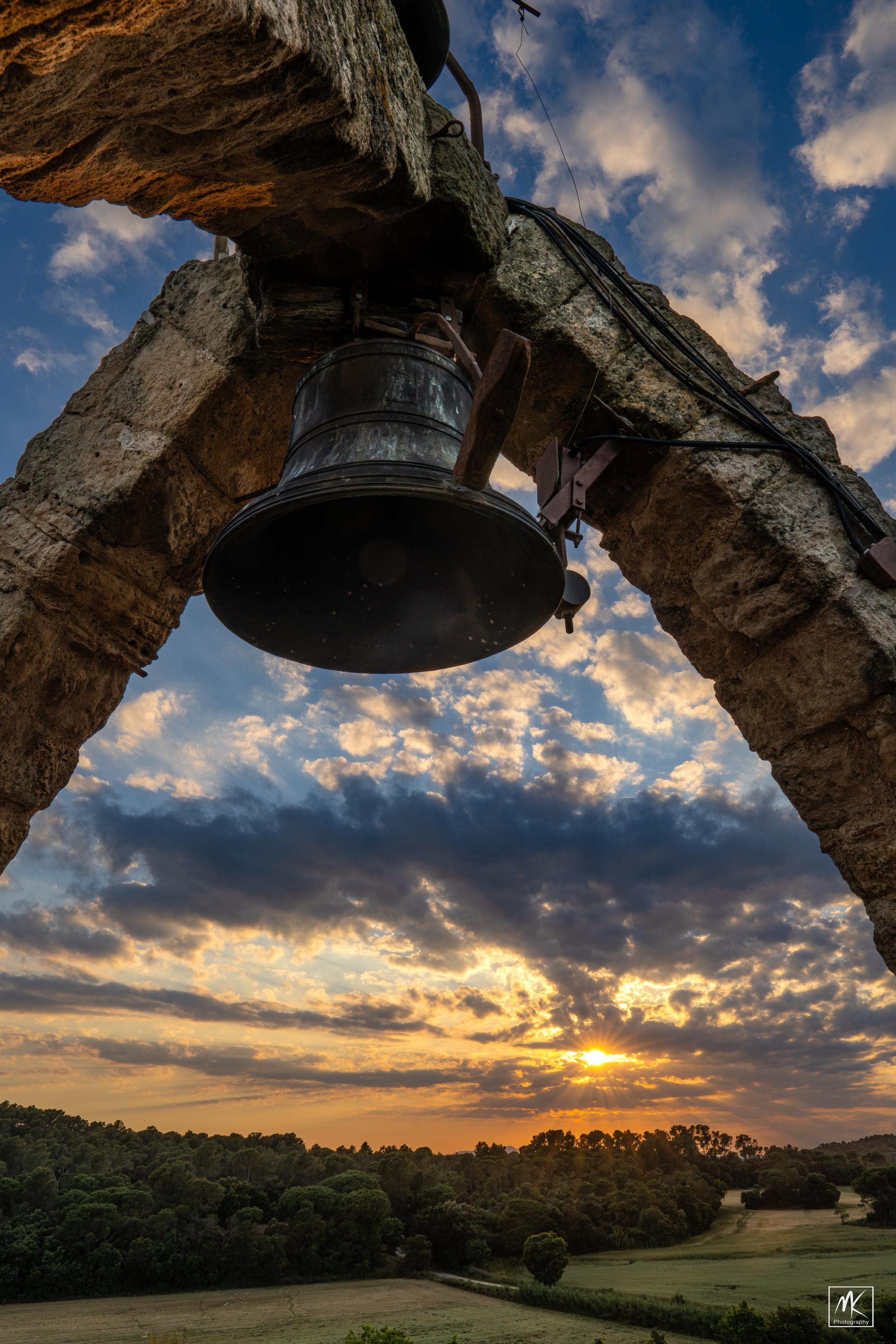 Color photo of a setting sun in a partly cloudy sky taken from the open top of a bell tower with the bell suspended overhead attached to the intersection of three curving stone arch supports.