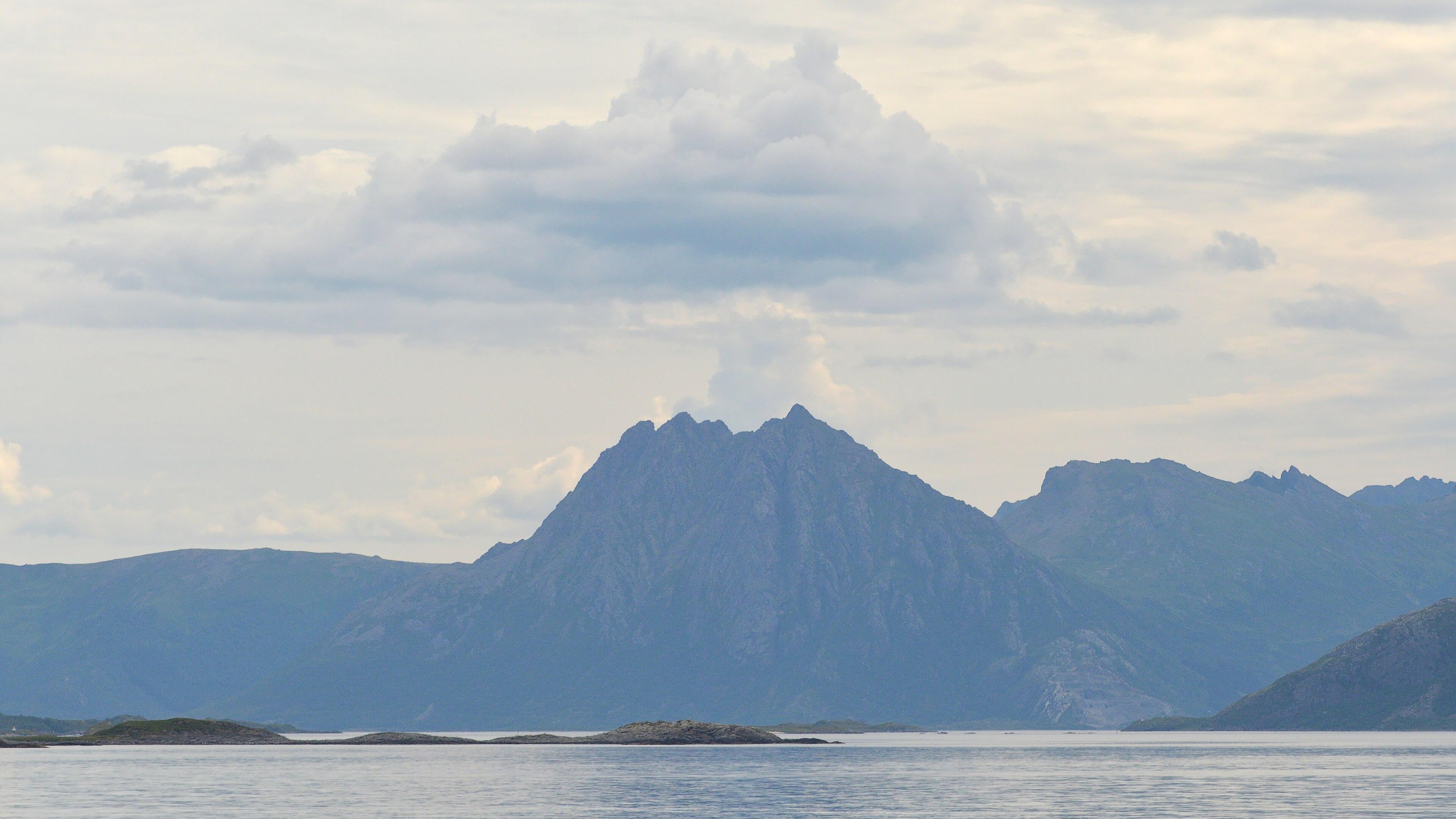 A photo of a mountain with water in front of it and clouds behind it that look like smoke from an erupting volcano.
