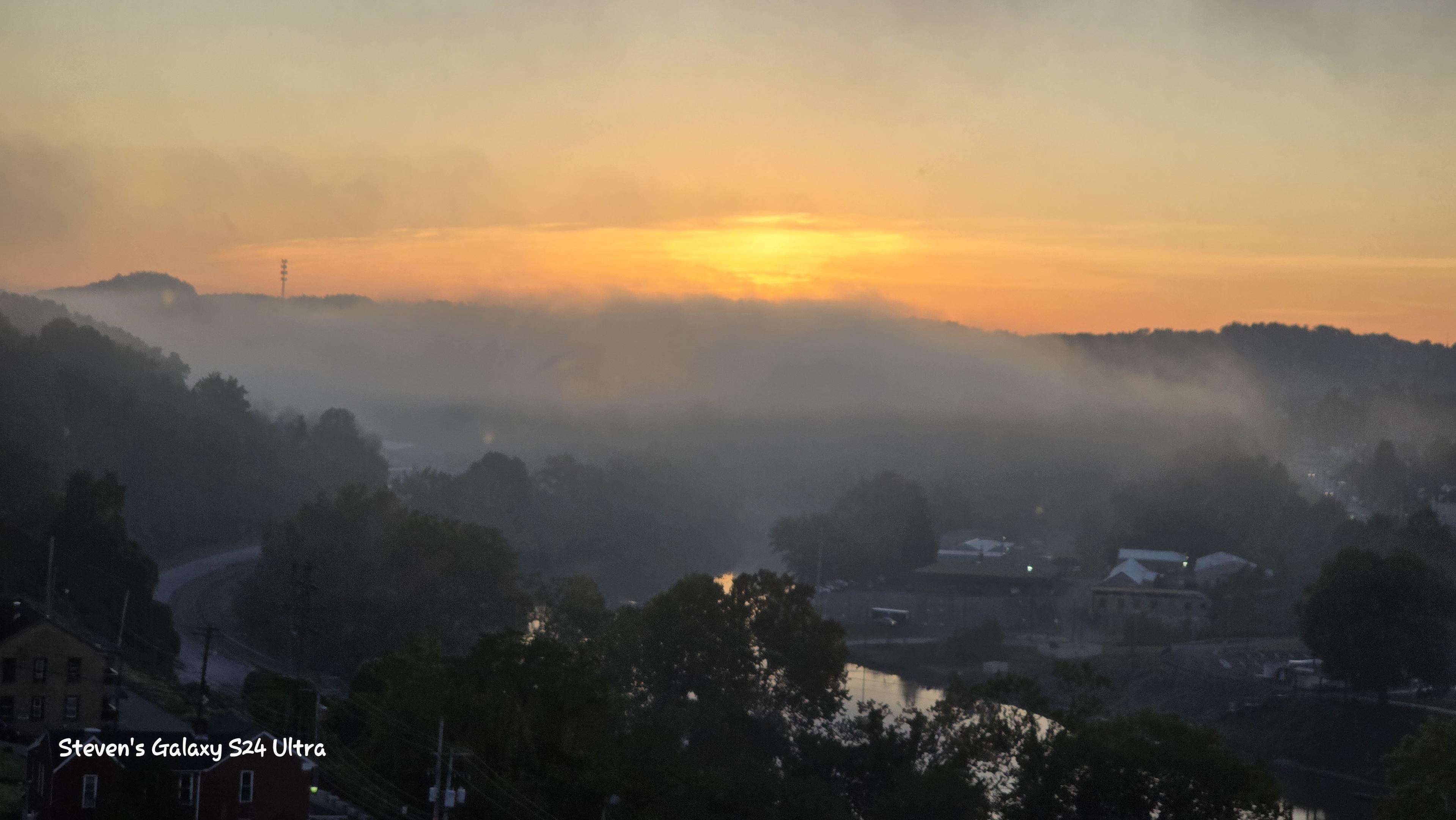 Misty sunrise over layered hills and a quiet valley, with dark trees, scattered homes, and golden light creating a peaceful mood.
