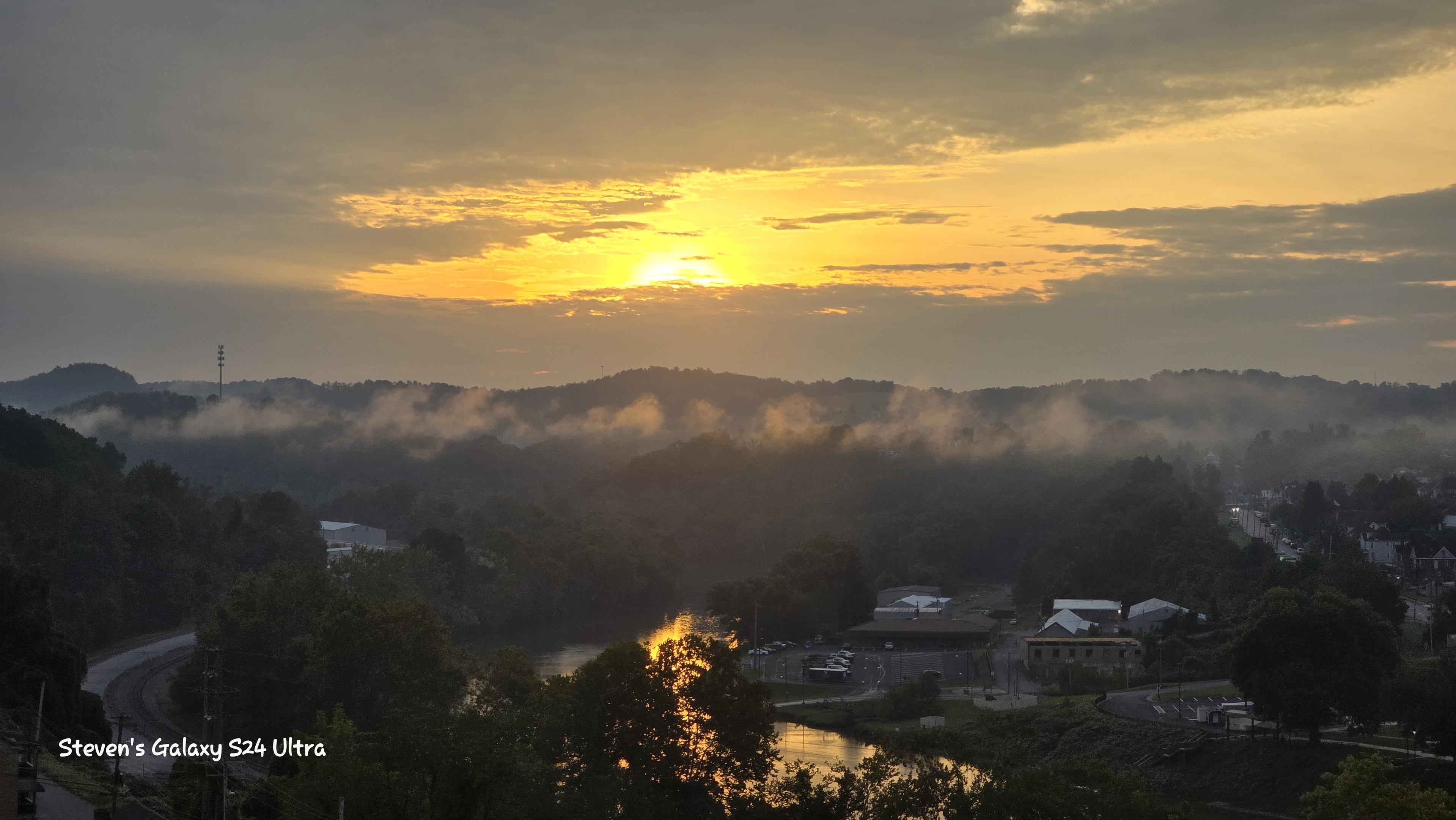Golden sunrise over misty hills and a quiet valley village below, with reflected light in a pond creating a peaceful, hopeful mood.