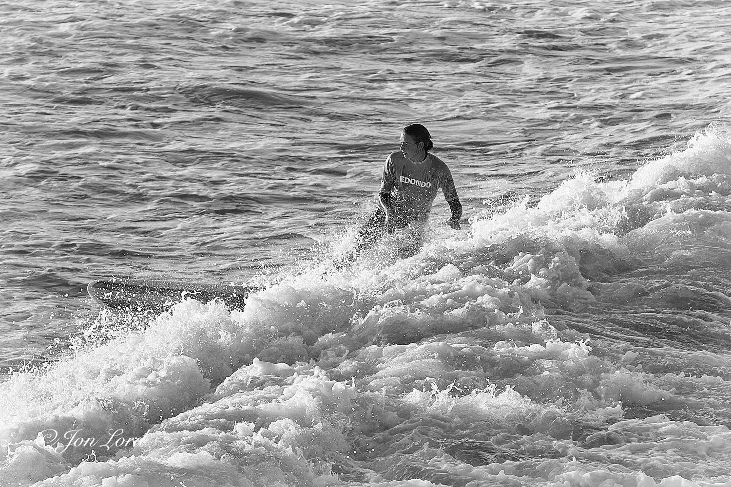 This is a black and white sports photo in landscape format of a young woman surfing at sunrise. Hermosa Beach, California (2013).

Stretching diagonally across the photo from the lower left corner towards the upper right is the foaming surf of a breaking wave that's moving from right to left. In the centre of the image is our teen surfer, surfing across the wave from right to left. She is viewed from about five metres away and two metres above and with her filling about half the height of the photo. Her body is facing the camera while her head is angled ahead, inline with the surfboard. She has dark hair that is tied behind her head, is wearing a light toned t-shirt with 'Redondo' printed on the front and covering her wetsuit. Below her waist is obscured by the breaking wave. Just visible ahead of the wave is the forward portion of her white surfboard that has two parallel black stripes inset from the edges. The sea ahead of the breaking wave is heavily disturbed with multiple small wa