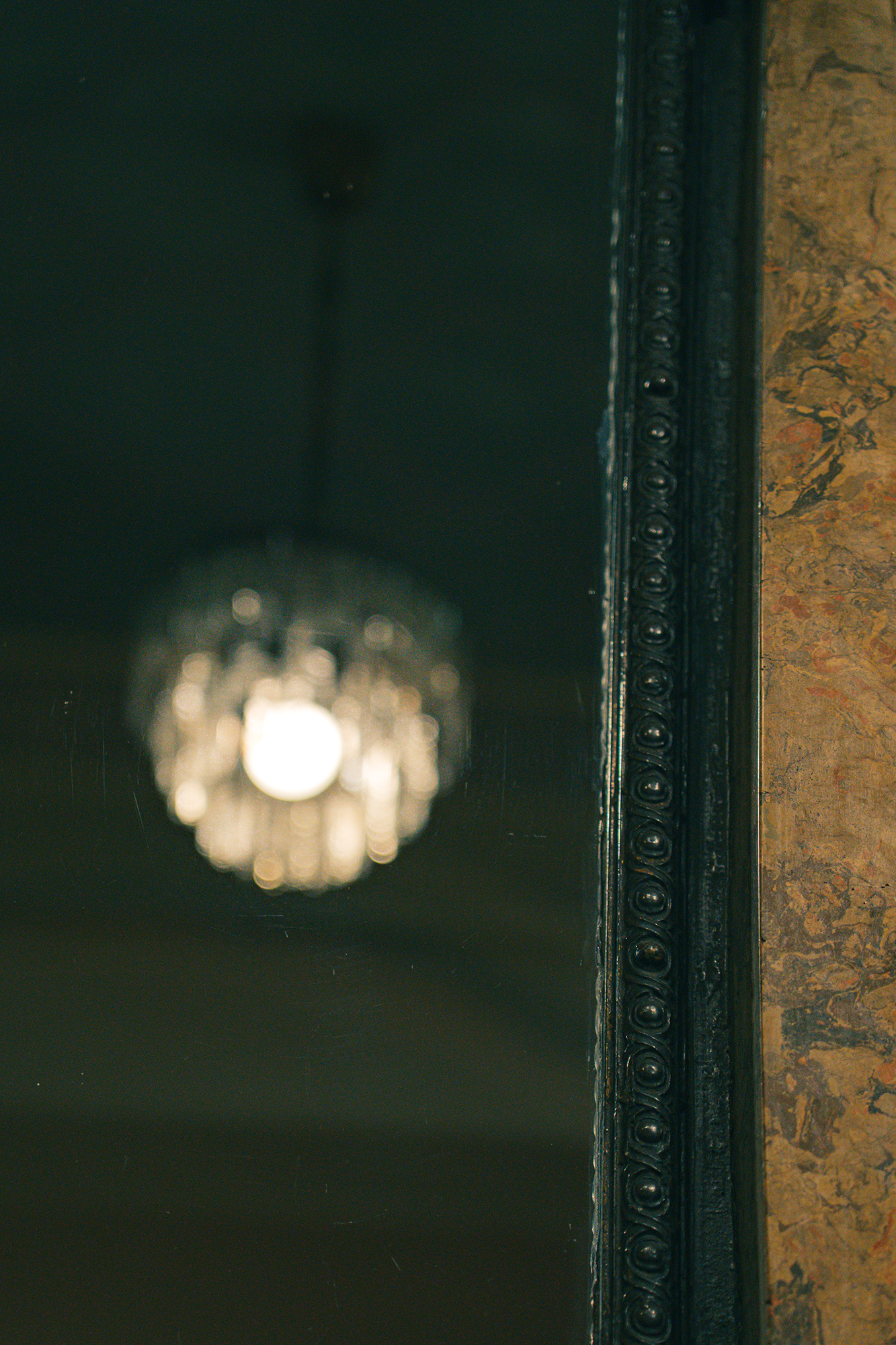 a photo of a blurry chanedlier reflected in an old mirror in a café. the mirror frame and the marble wall next to it are in focus