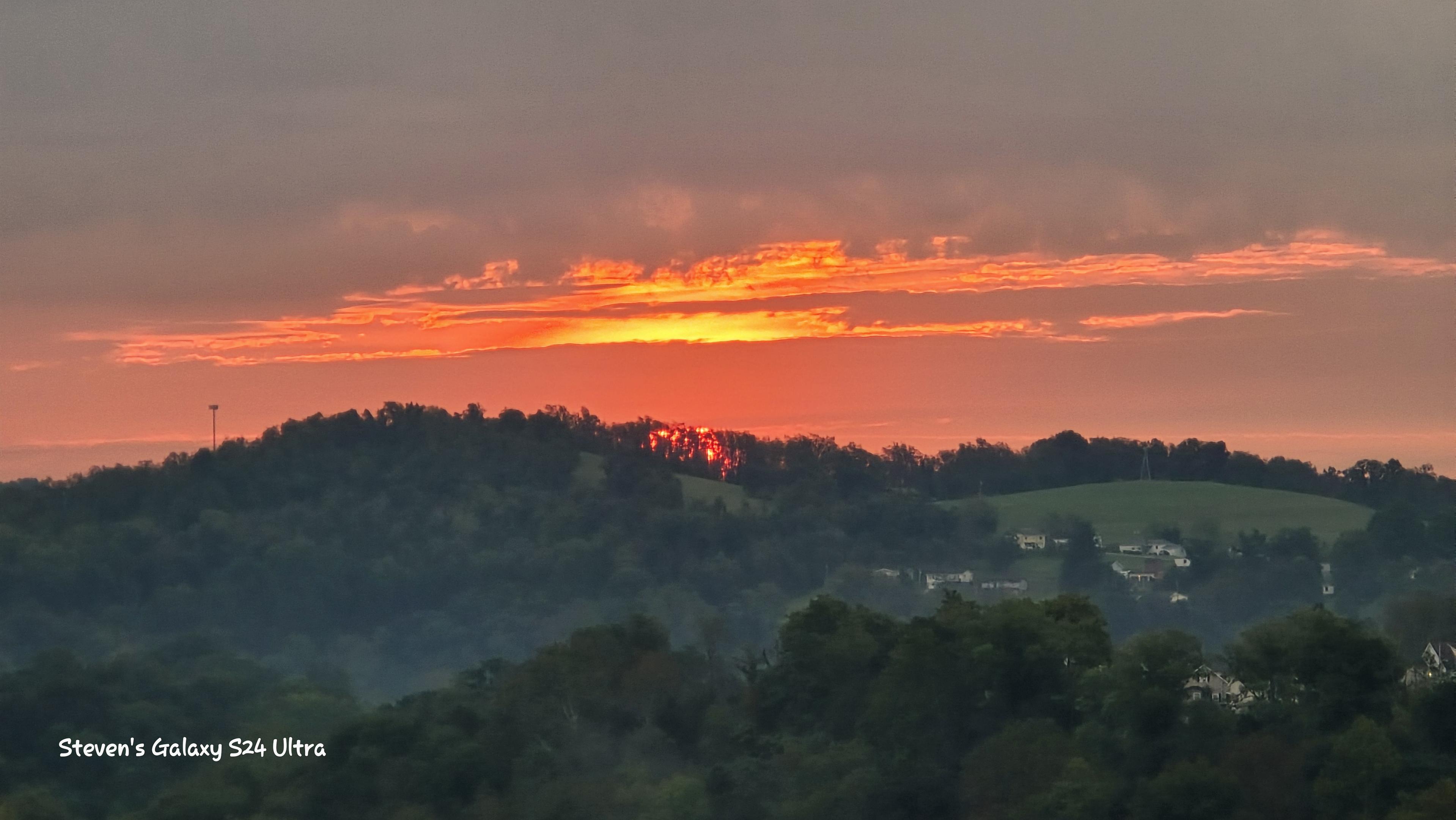 Fiery orange sunset glows beneath gray clouds over misty, tree-covered rolling hills and scattered homes, creating a peaceful rural scene.