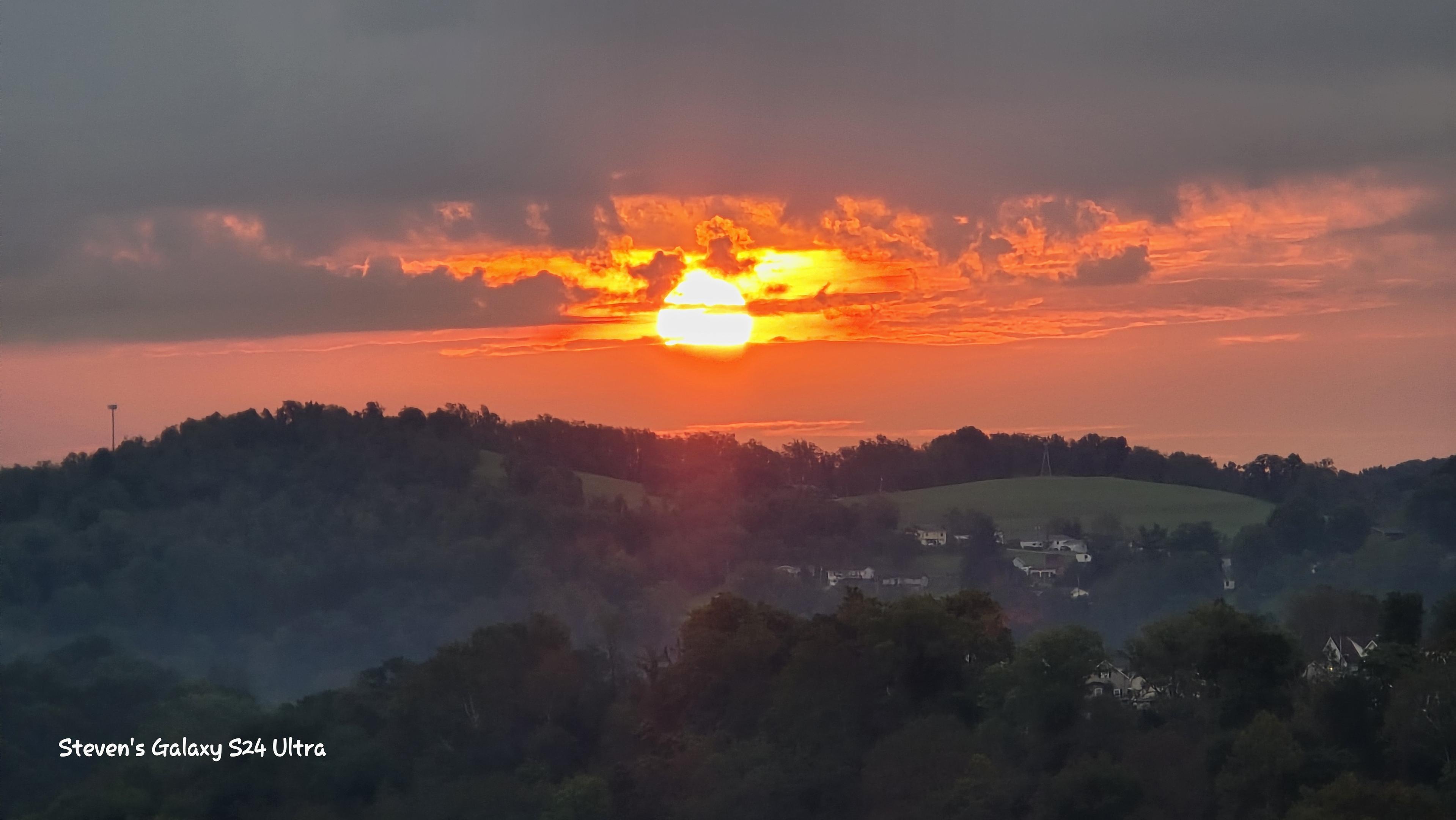 Fiery orange sunset breaks through dark clouds above forested rolling hills and scattered homes, casting a warm, dramatic glow.