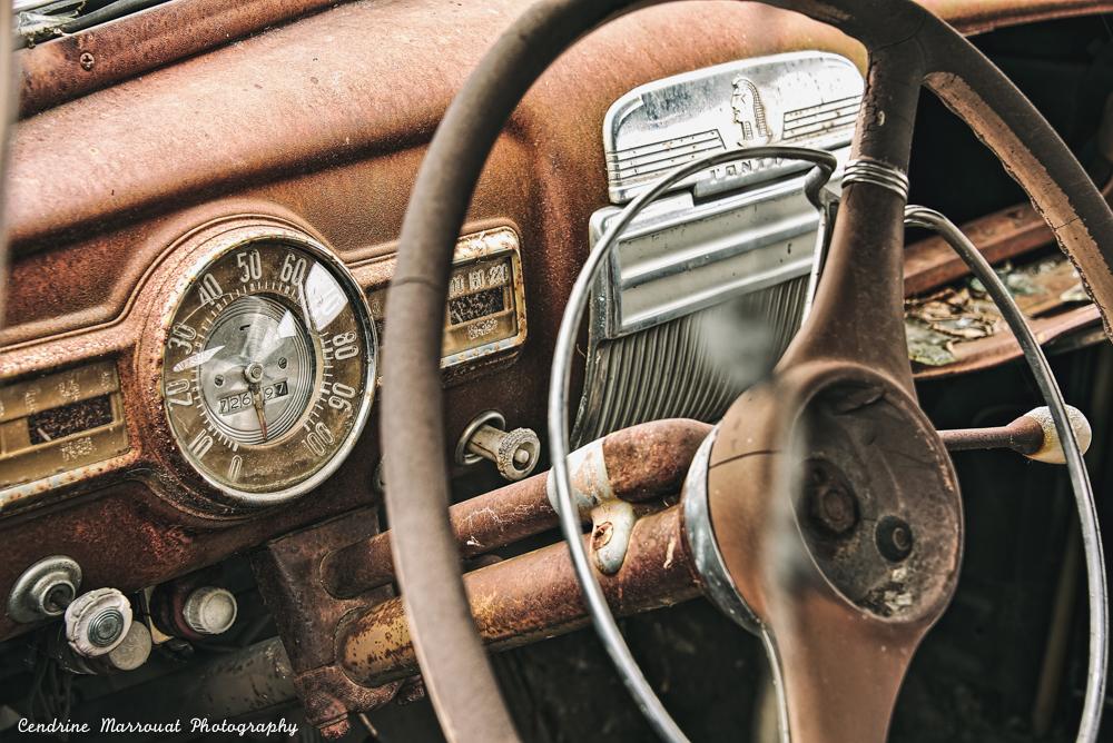 The dashboard of an old car, probably from the 1950s or 1960s. It is rusty and the spinner knob of the steering wheel is missing.