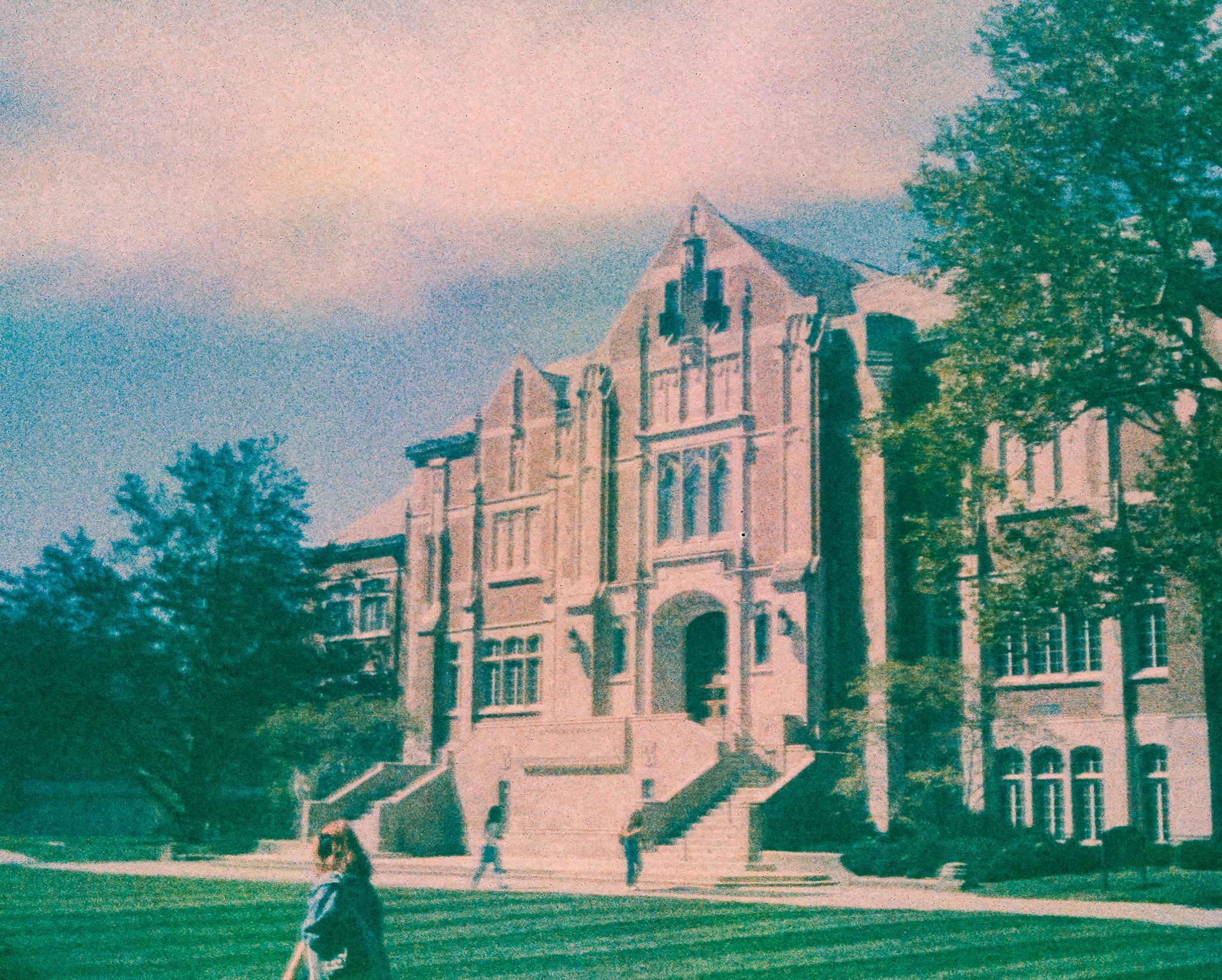 An ornate brick building with many windows, pointed roofs, a large arched doorway, and a large double staircase. There are trees to the left and right of the building. There is a concrete walkway in front of the building, and two people are walking past the front of the building. There is a green lawn in the foreground, and a person walking to the left, not far from the camera. The sky is partly cloudy and partly blue. The photo is very grainy, and the colors are muted, with a somewhat pinkish cast.
