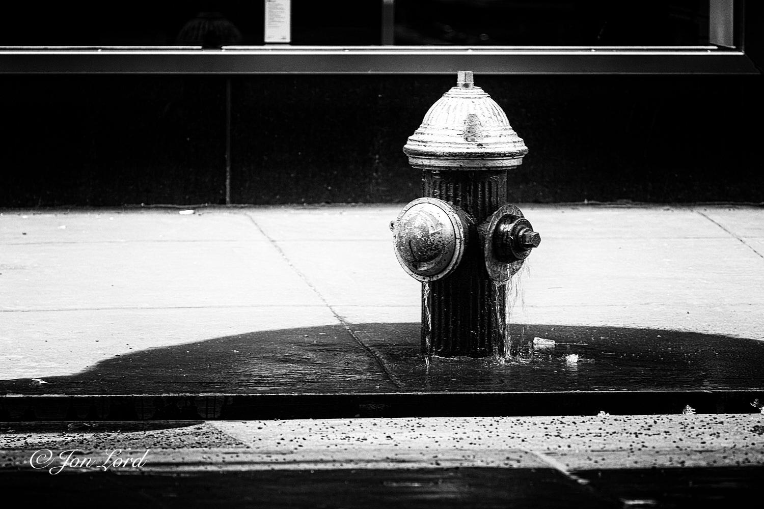 This is a grungy black and white street photo in landscape format of a leaking fire hydrant on the side of a city pavement with a few small chunks of ice to one side. Madison Ave, NYC (2012).

In the foreground is the road surface rising up from the base of the photo to less than the quarter way up level. At the base, the surface is jet black, then becoming a lighter tone closer to the metal curb strip where the pavement start. Slightly to the right of centre is our black cylindrical fire hydrant. The hydrant is viewed from the same level and from two metres away. It rise up a little over one metre with a silver cone shaped top, peaked by a large square shaped nut. Below the cone, on the left side is a dome shaped cover with a hexagonal nut on the right side. From both the dome and the nut is a steady stream of water falling a short distance onto the pavement below. Surrounding the hydrant is a semicircle, the straight edge is formed by the curb with the curve arcing around behind the 