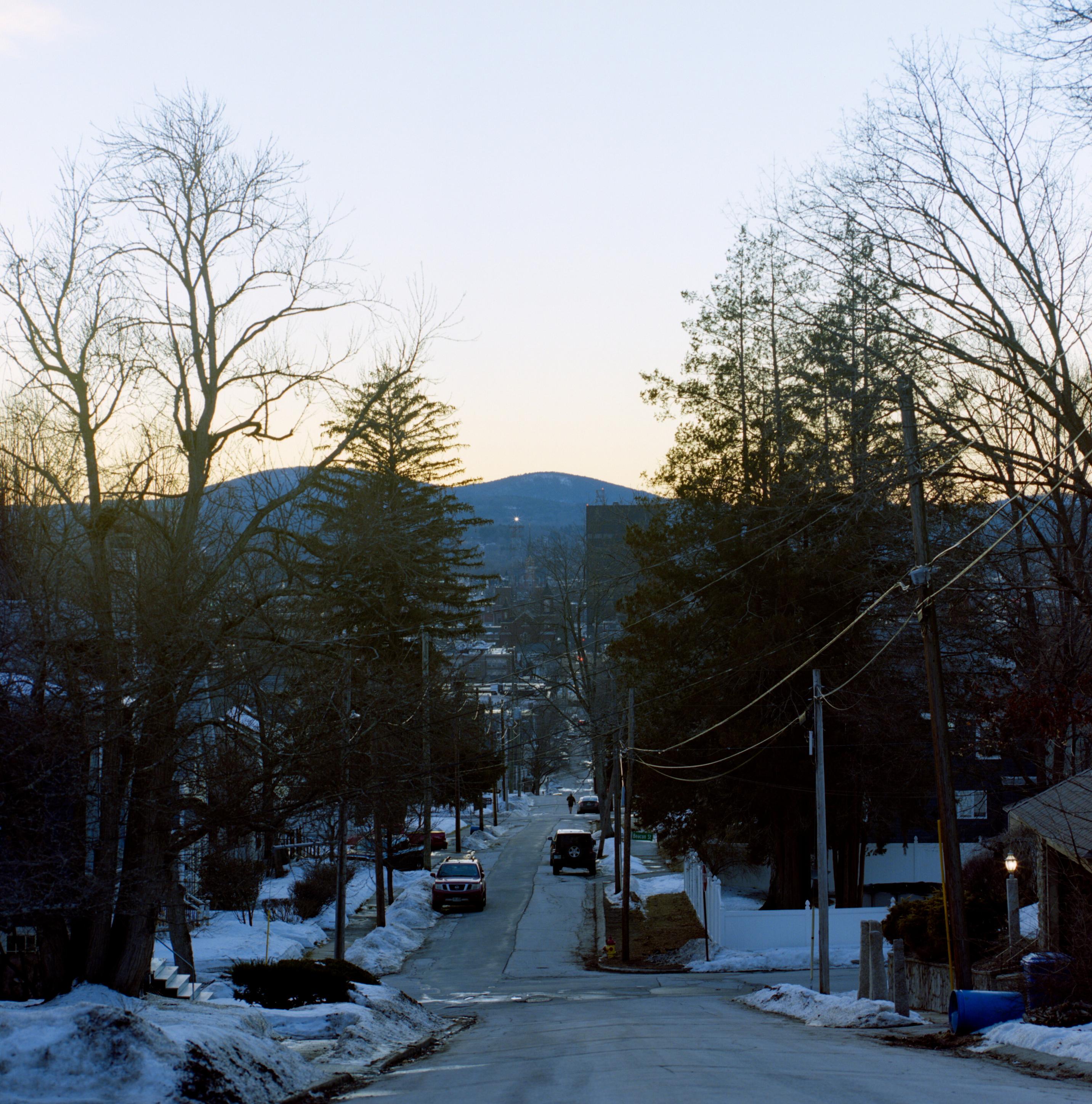 an image of a winter street stretching into the distance, where a pair of hills stand. bare trees accent the edge of the street, and a few cars are parked outside the residences of the small city.