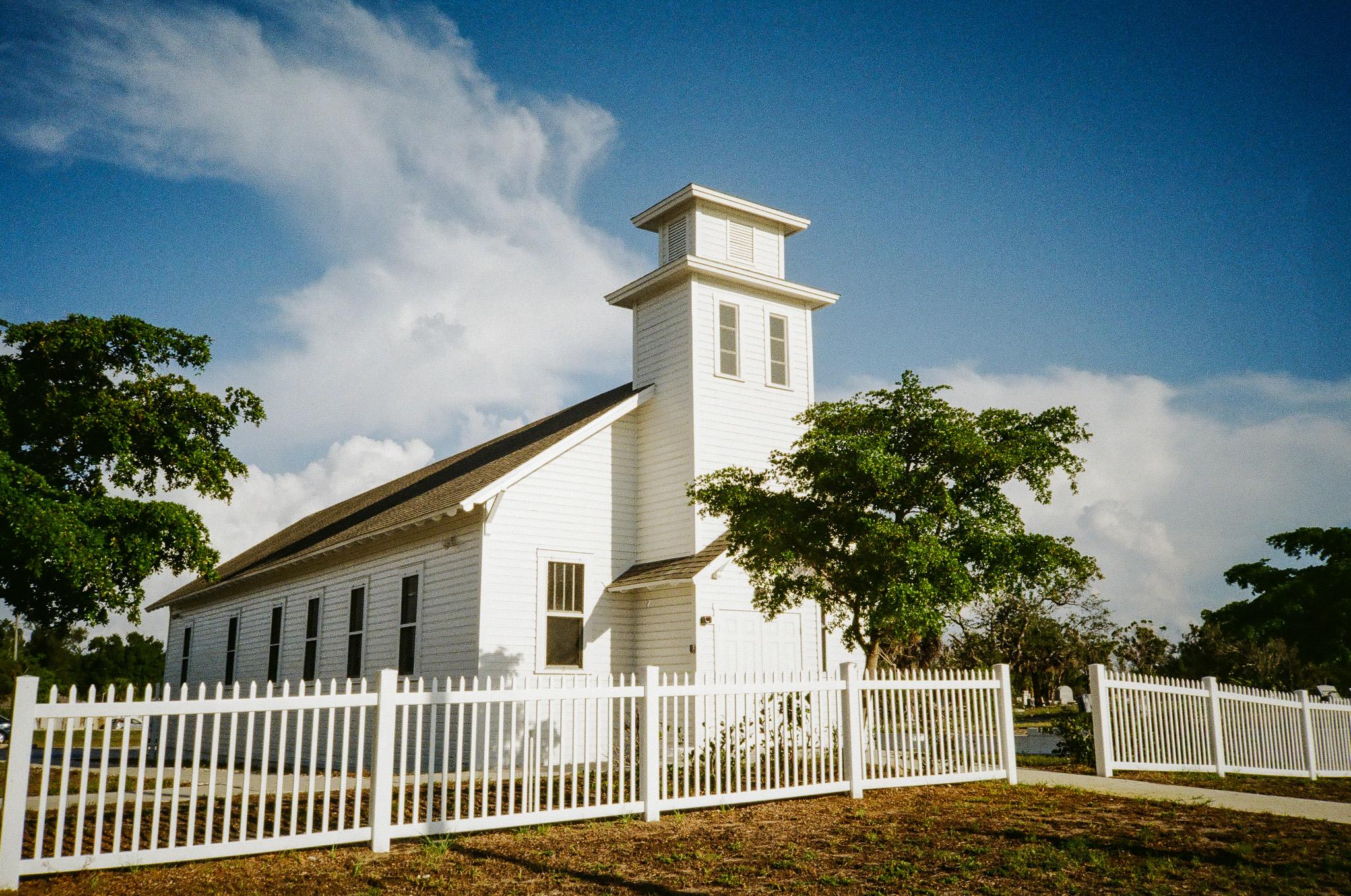 A traditional white church building with a sloped roof and windows in the front and along the side walls. In the front there is a tower extending above the roof, but there is no pointed steeple at the top. There are two windows near the top of the tower. There is a white picket fence along the front of the church, and a paved walkway leading to the front door. The ground is brown with small bits of green grass or weeds spaced apart. There are green trees to the left, in front of the church, and to the right. In the distance to the right, small gravestones can be seen. The sky is a dramatic blue color, with white clouds above the horizon and extending above the church and to the left.