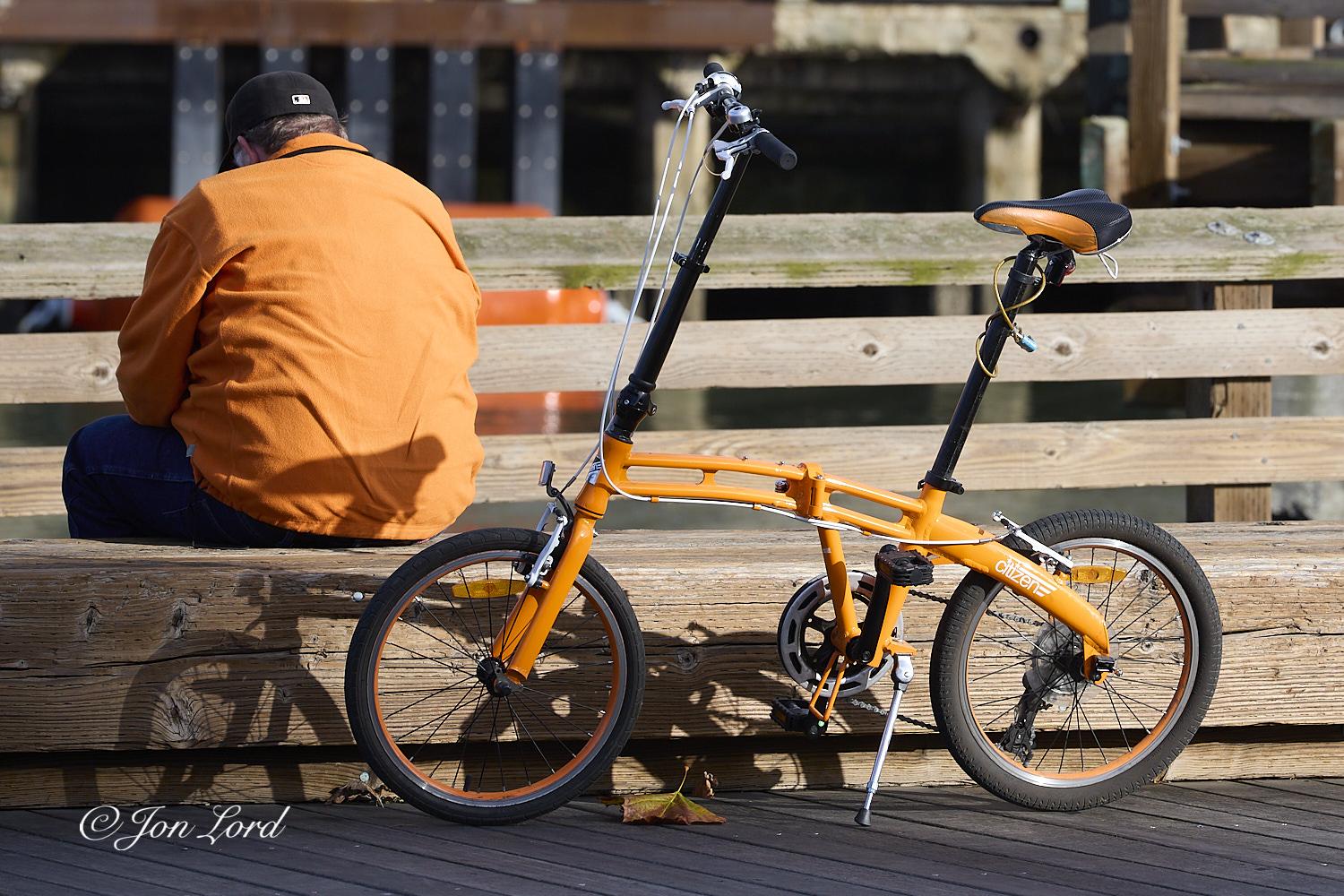 This is a colour street photo in landscape format of a man wearing an orange jacket sitting on a bench next to an matching orange bike. Pier 35, San Francisco (2012).

In the foreground the ground is made up of neatly laid wooden planks extending away and diagonally from the camera which is located half a metre above the ground. Two metres from the camera is a light brown wood, long and heavy section with a square cross section of half a metre that stretches across the photo from the left, to the right edge and is purposed as a bench. Against this stout length of timber and in the centre of the image, leans the orange bike, the bike and most of the scene is brightly lit from the sun that's behind the camera. The cycle appears to be collapsable with small, half a metre sized wheels, a low set frame with handle bars mounted on a long black tube with the saddle fixed in a similar fashion. The rims of the tyres, frame, peddles and saddle are all a caramel shade of orange. To the left of ce